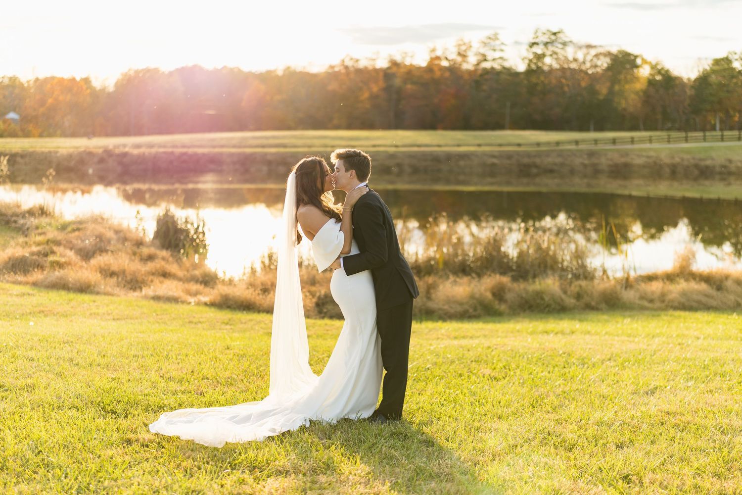 Bride and groom sharing romantic kiss at golden sunset beside serene pond at outdoor wedding.