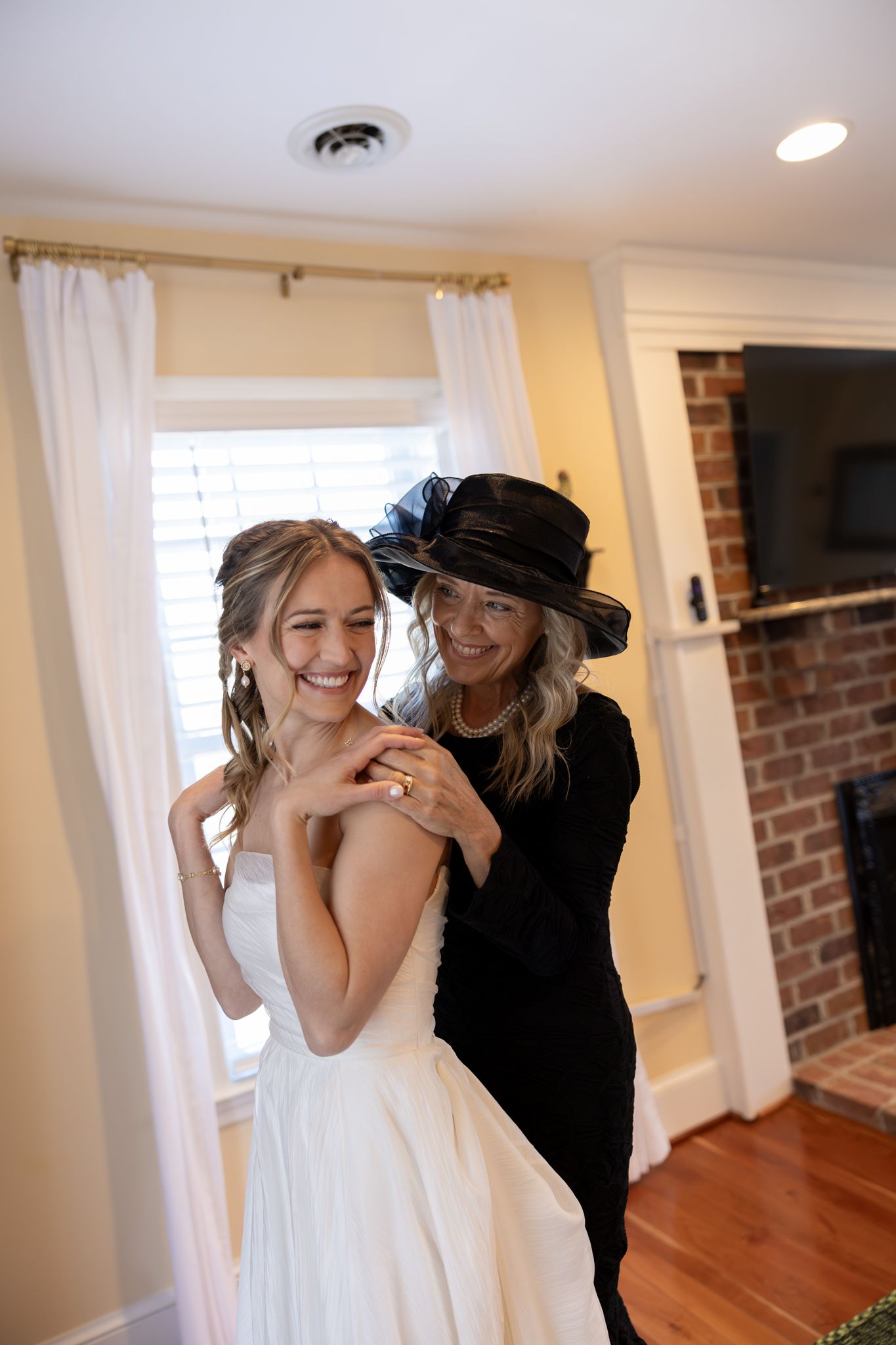 Bride smiling as an older woman in a black hat lovingly places a necklace on her in a bedroom.