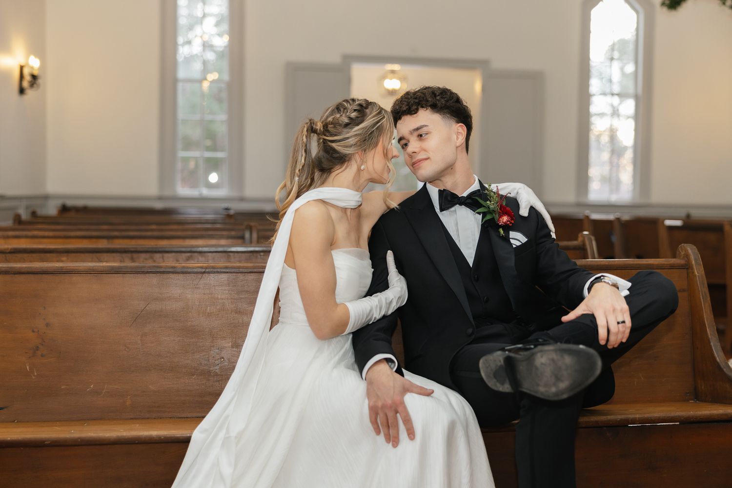 Elegant bride and groom share an intimate glance while seated in wooden church pews during wedding.