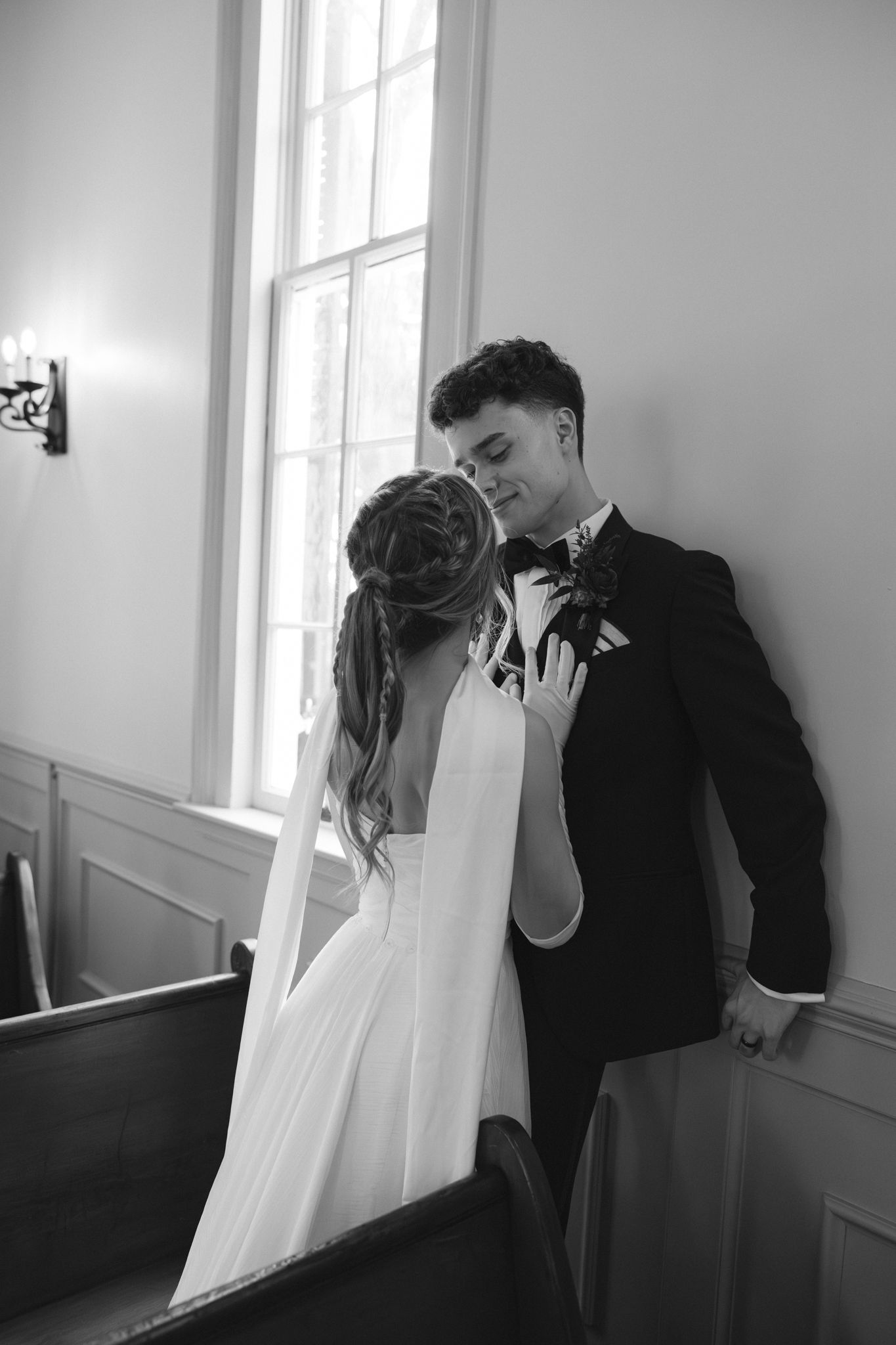 Black and white portrait of bride and groom sharing intimate moment by church window in chapel pew.