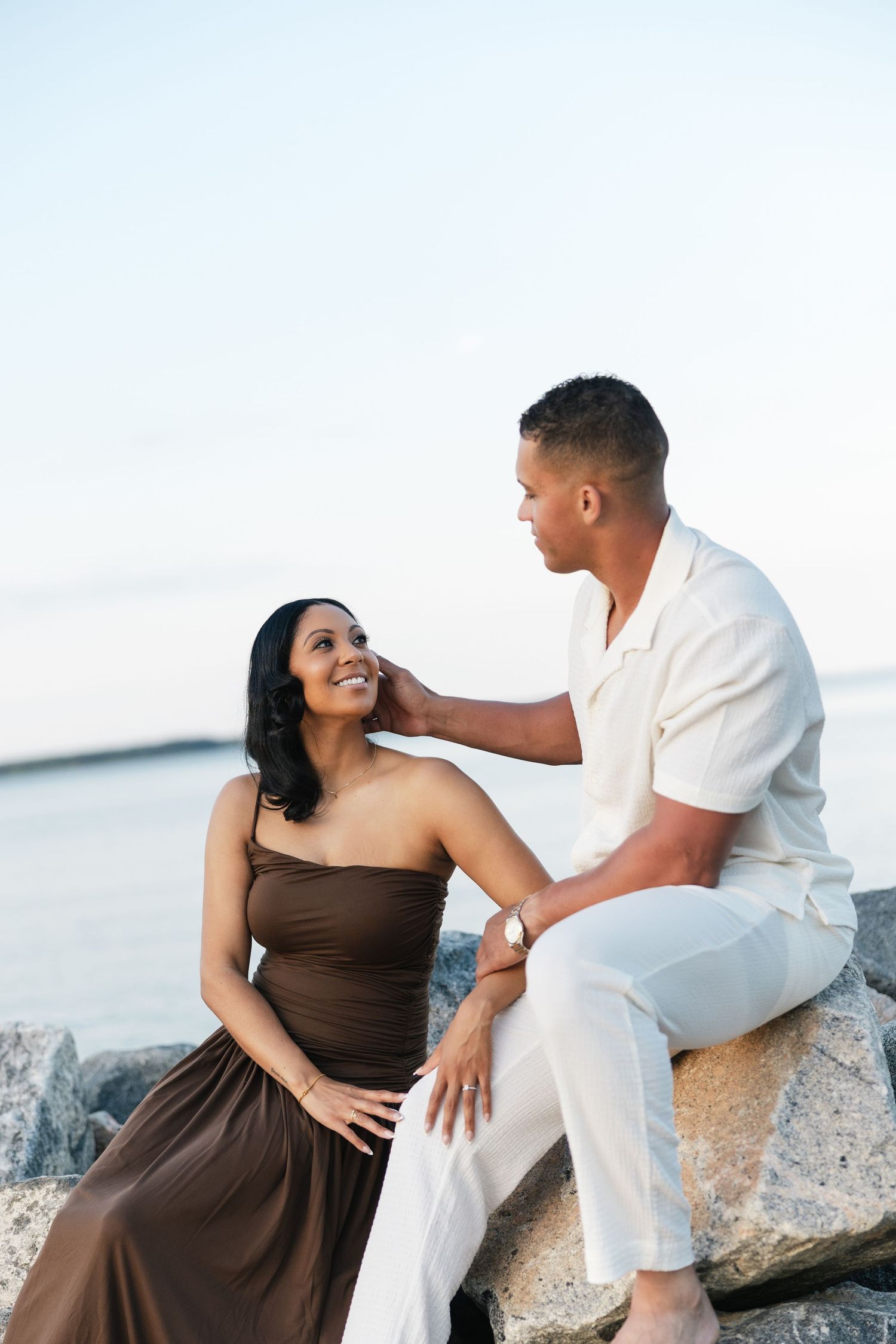 A couple sitting on rocks by the ocean, man in white outfit gently touching woman's face in brown dress.