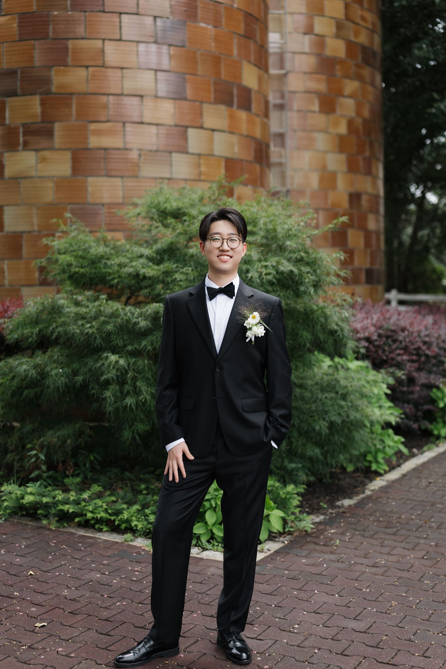 Young man in black tuxedo with boutonniere and glasses smiling outdoors near brick wall and green shrubs.