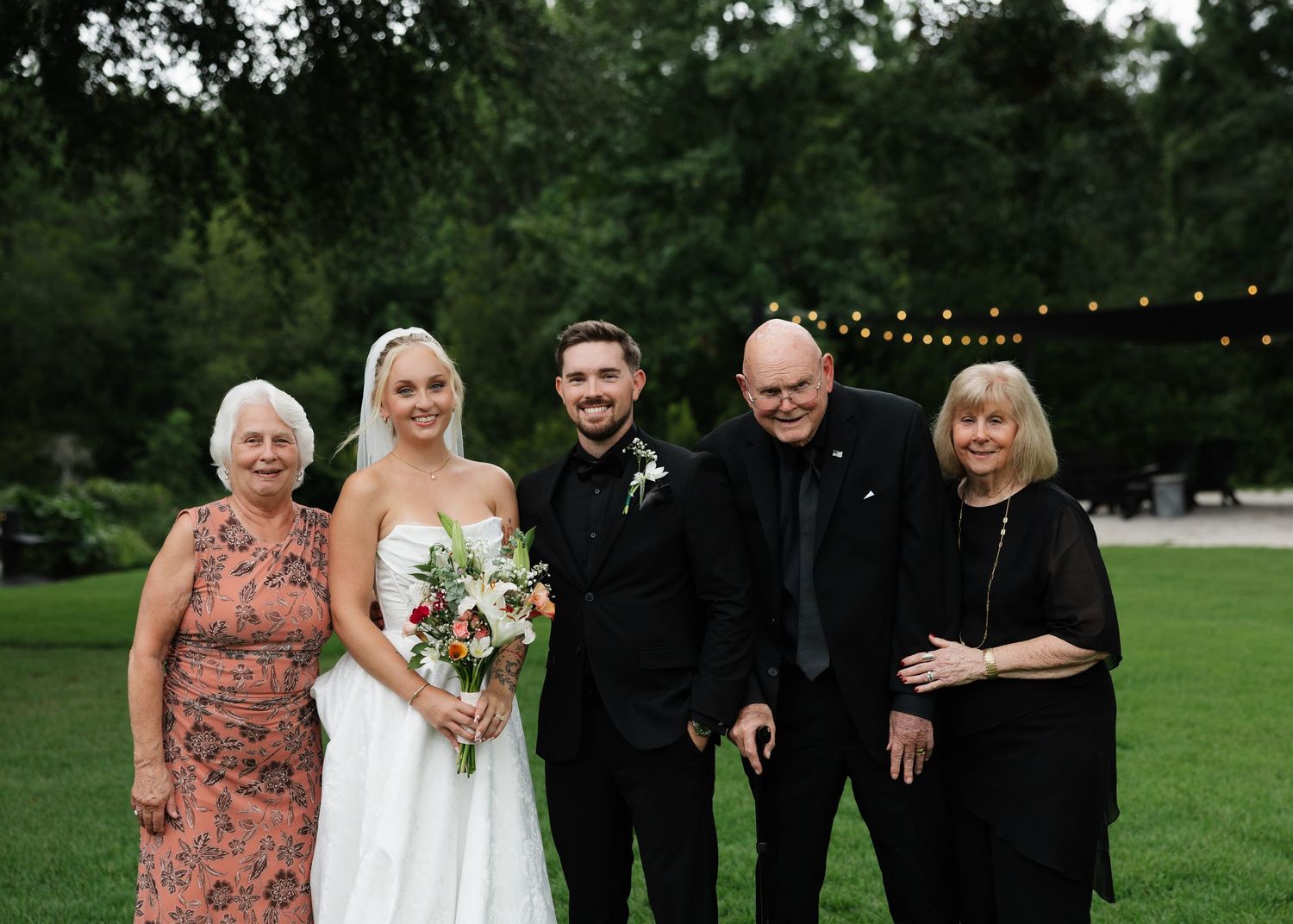 Wedding party of five posing outdoors on green lawn with string lights and trees in background.