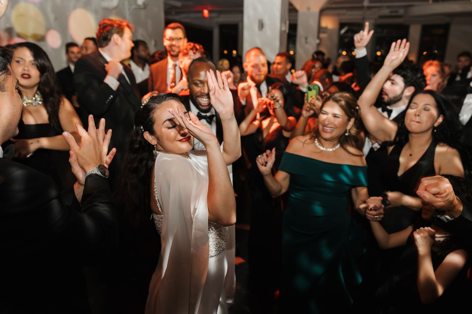 Guests dancing joyfully at a wedding reception, with the bride in a silver gown raising her hand on the dance floor.