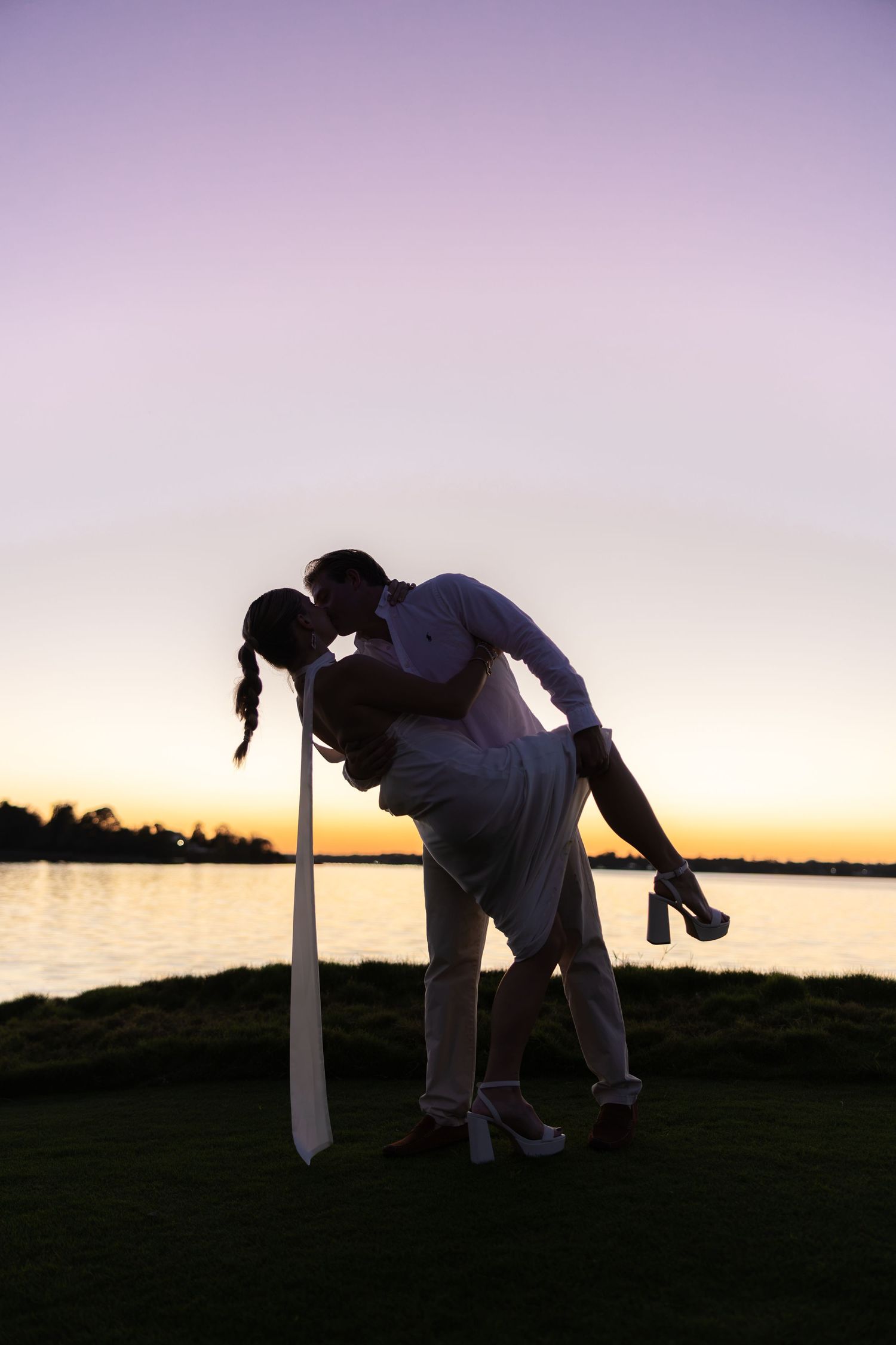 Couple sharing a romantic dip kiss silhouetted against a stunning purple sunset by the waterfront.