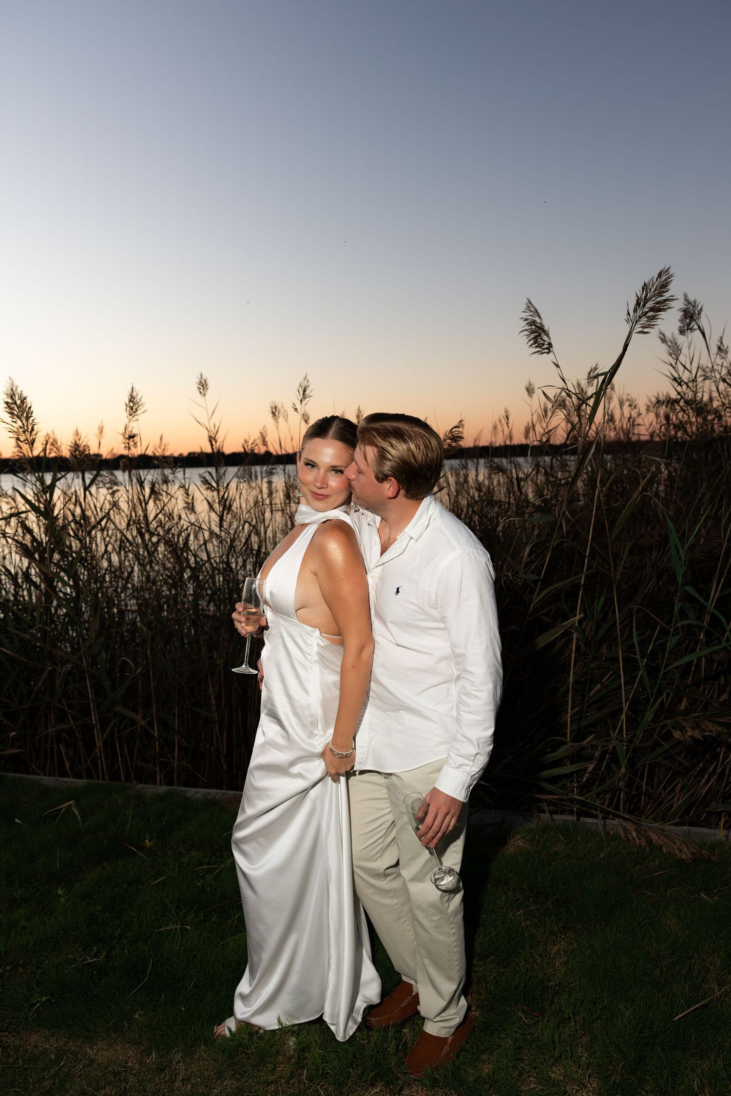 Couple in white attire sharing a kiss at sunset near a waterfront with tall reeds in the background.