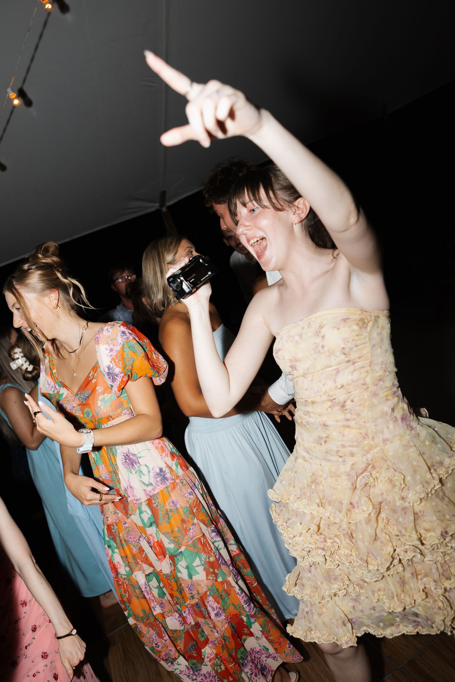 Young woman in ruffled dress dancing joyfully at a party, pointing up, surrounded by guests in colorful attire.