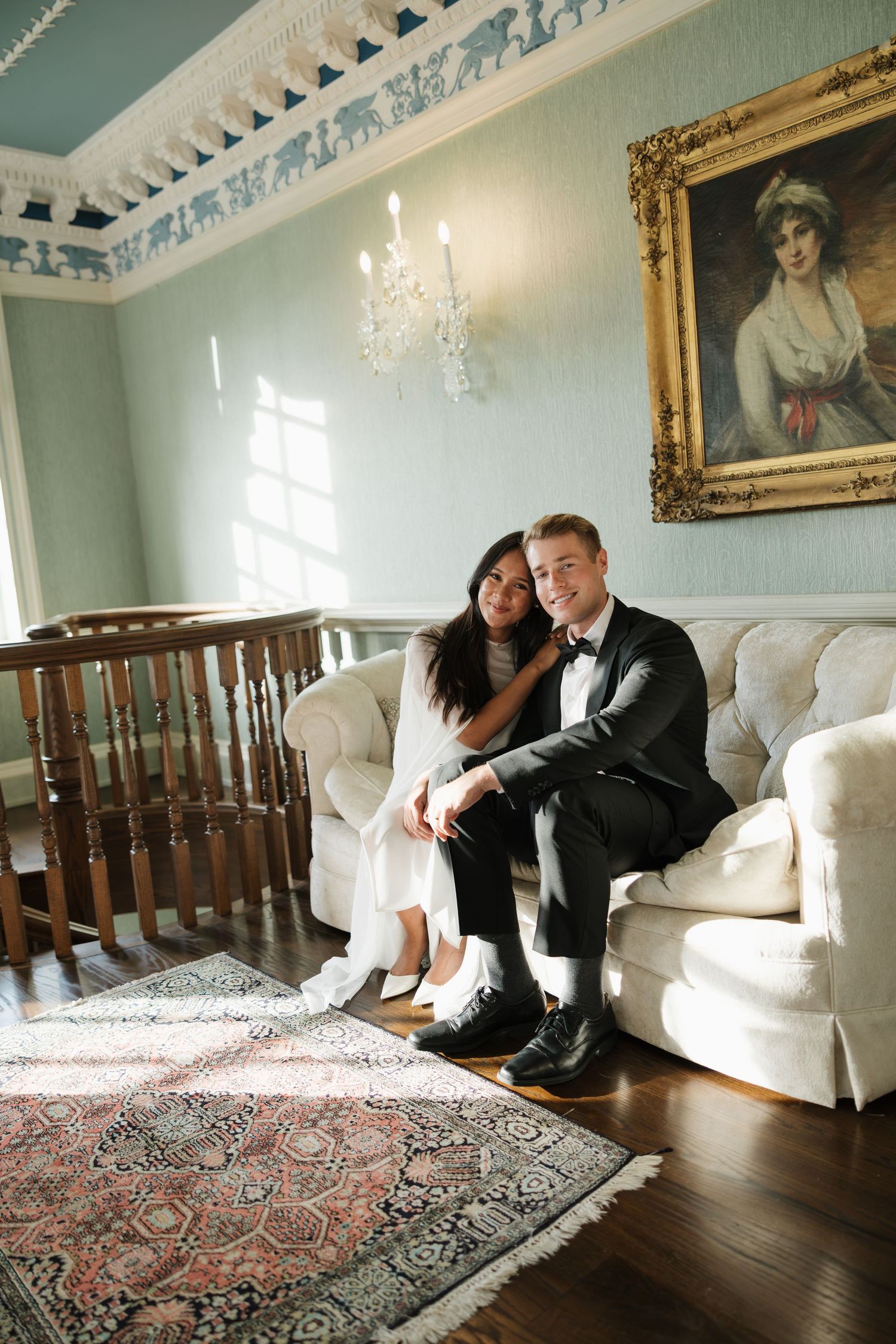Elegant couple in wedding attire sitting on a white sofa in a classic room with ornate decor and portrait painting.