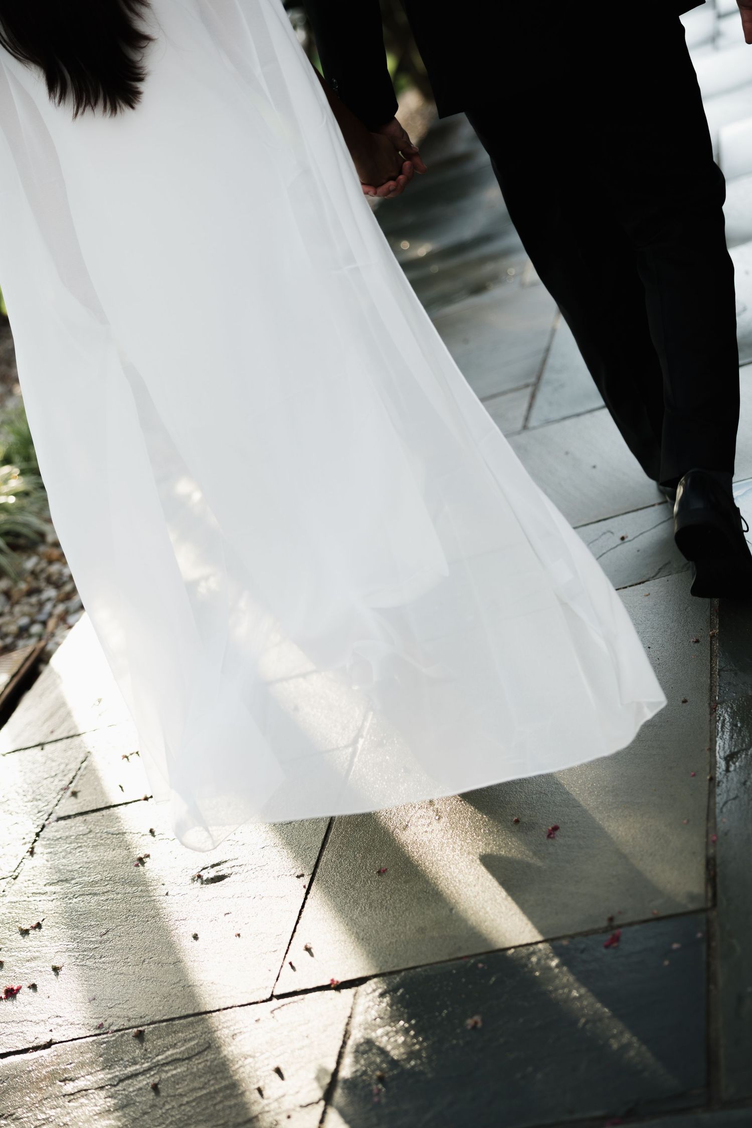 Bride in flowing white wedding gown walks with groom in black suit on stone pathway, veil billowing in the breeze.