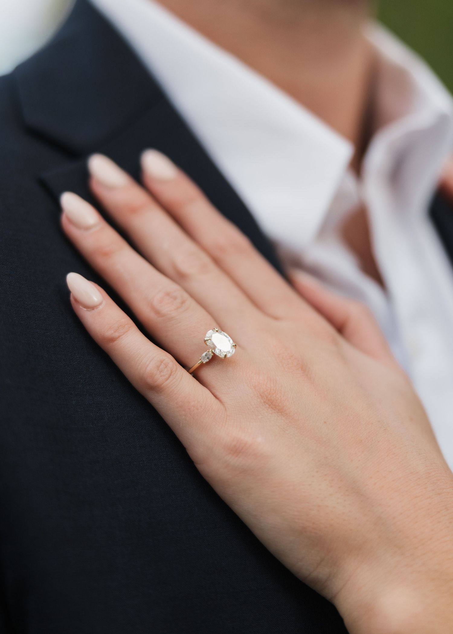 Close-up of a diamond solitaire engagement ring on a woman's hand resting on a man's dark suit jacket.
