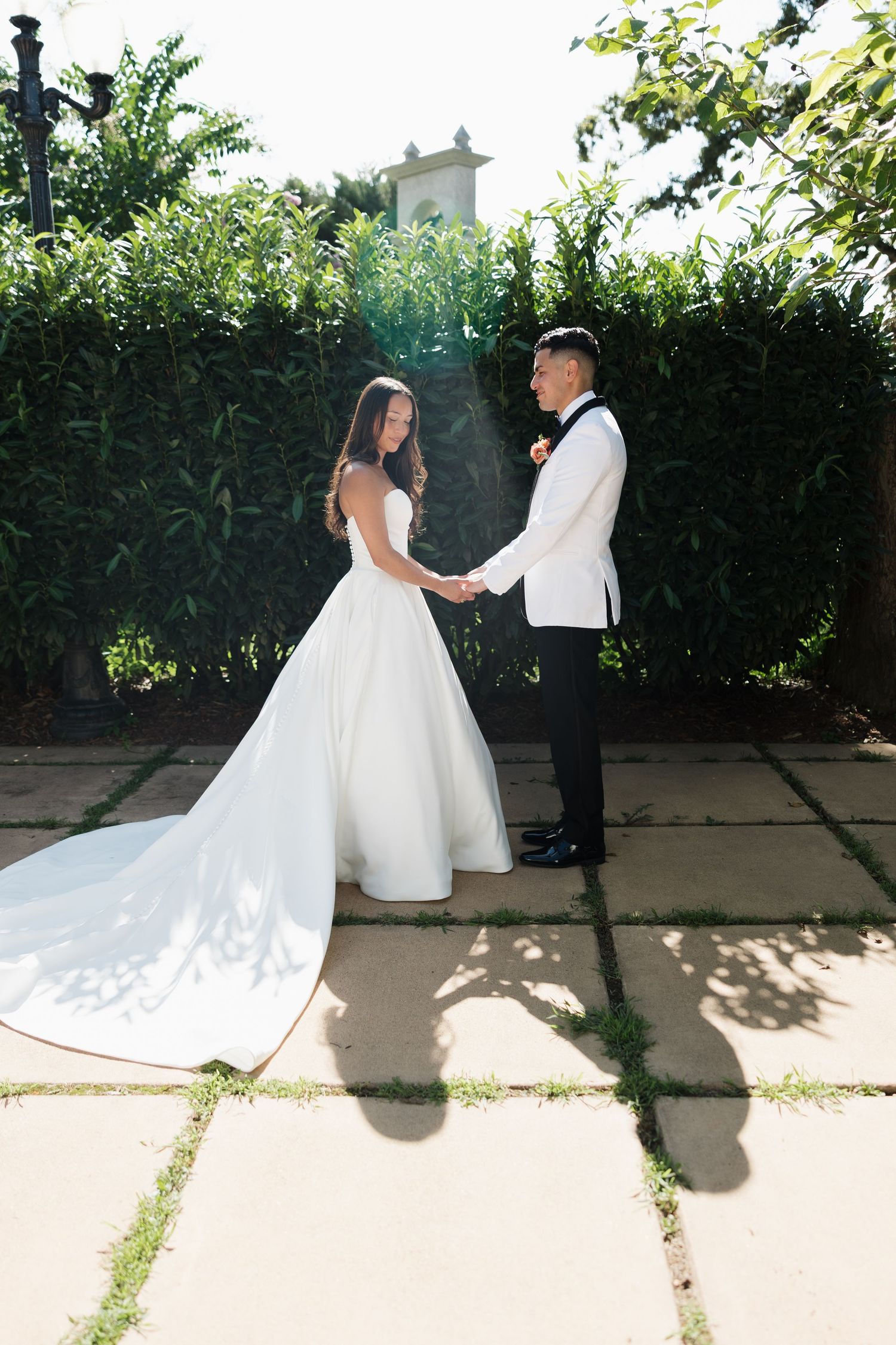 Bride and groom holding hands in garden, bride wearing elegant white gown with long train, groom in white tuxedo.