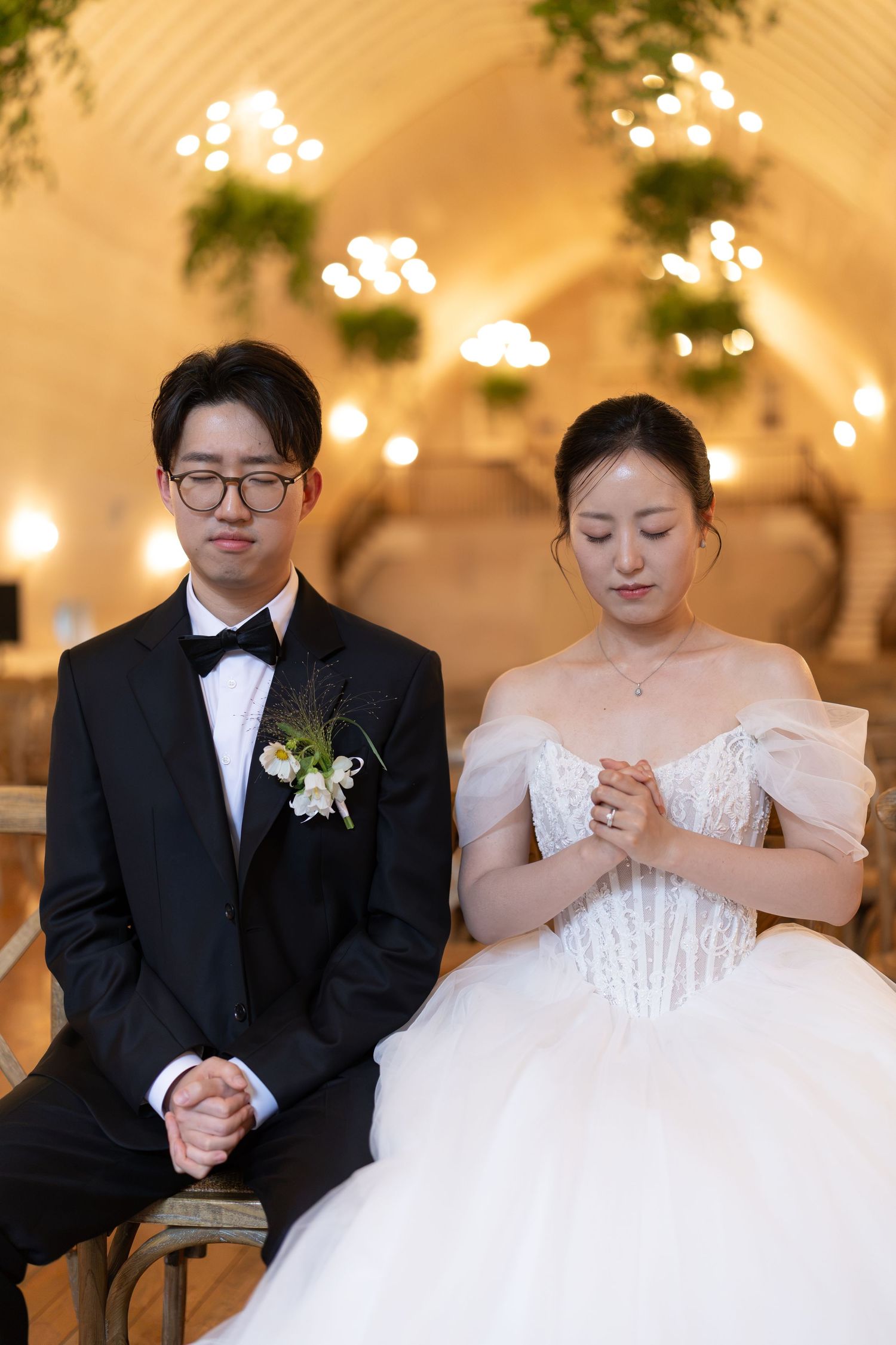 Asian bride and groom sit together in prayer during their elegant candlelit wedding ceremony indoors.