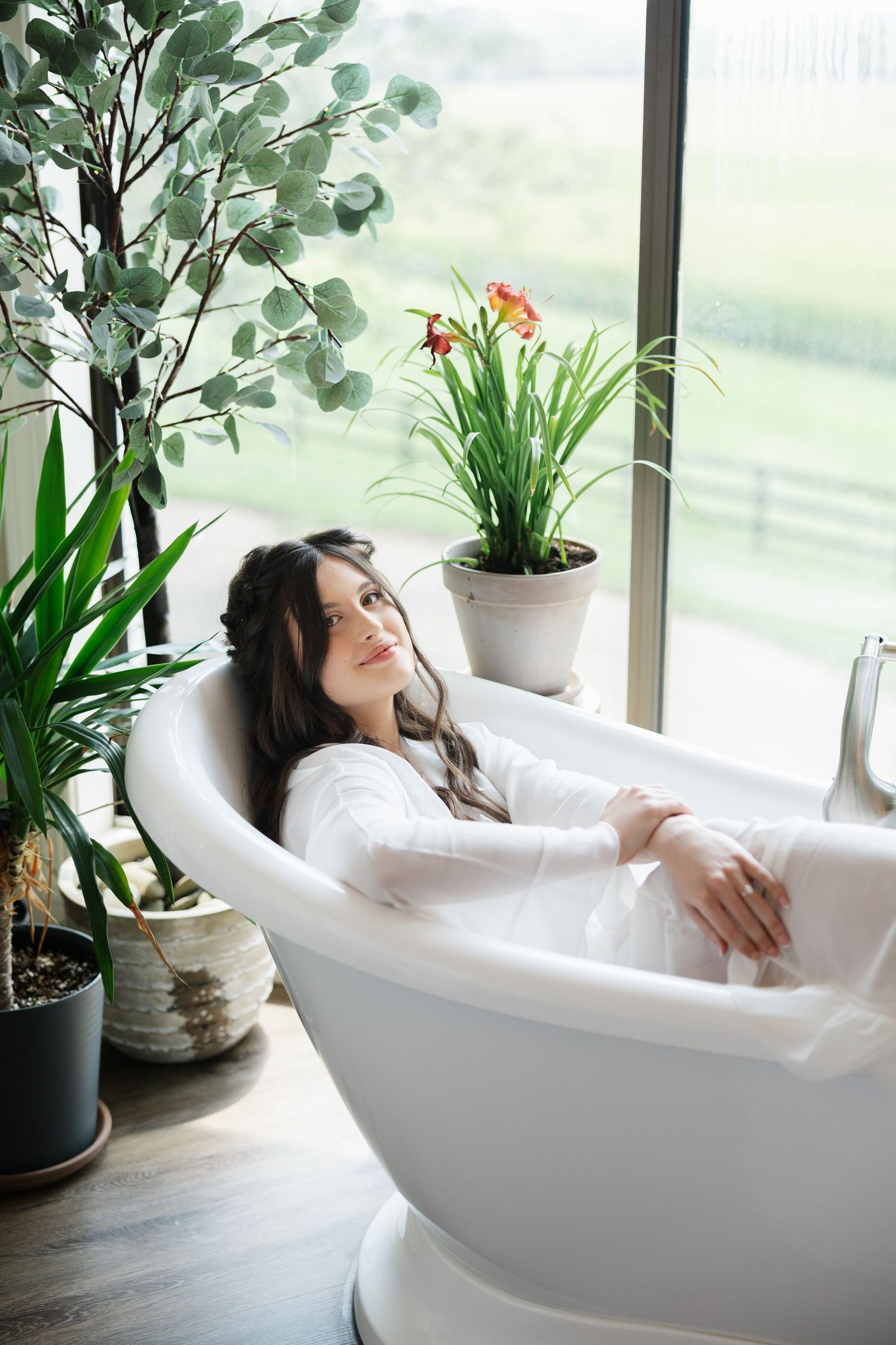 Woman relaxing in a freestanding white bathtub surrounded by lush green plants near a large window.