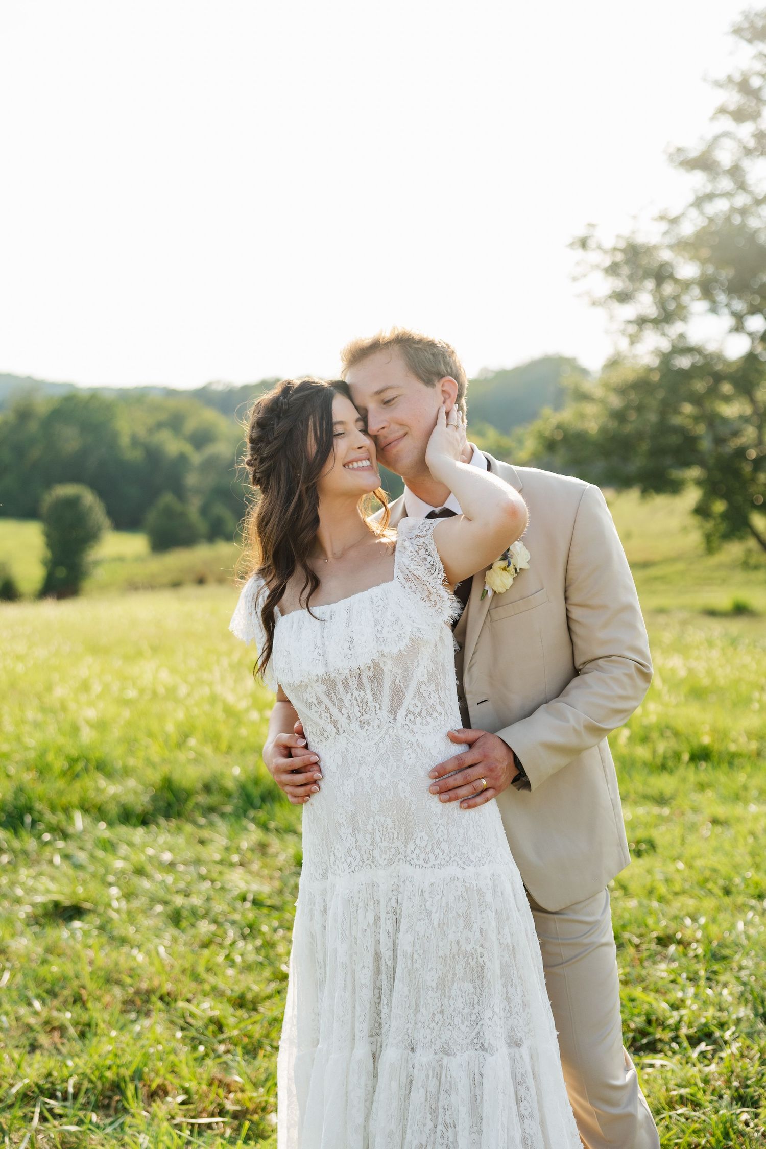 A bride in white lace dress and groom in tan suit share a romantic moment in a sunny green meadow.