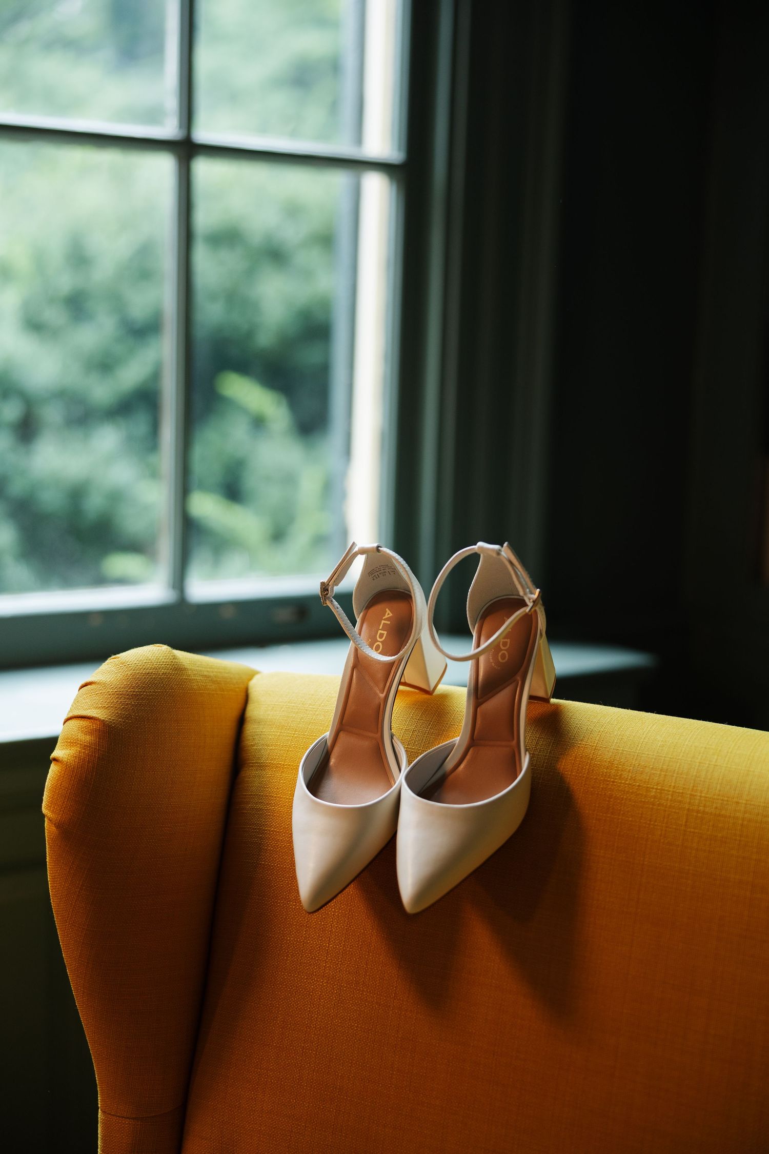 Elegant white pointed-toe heels with ankle straps resting on a yellow chair near a window.