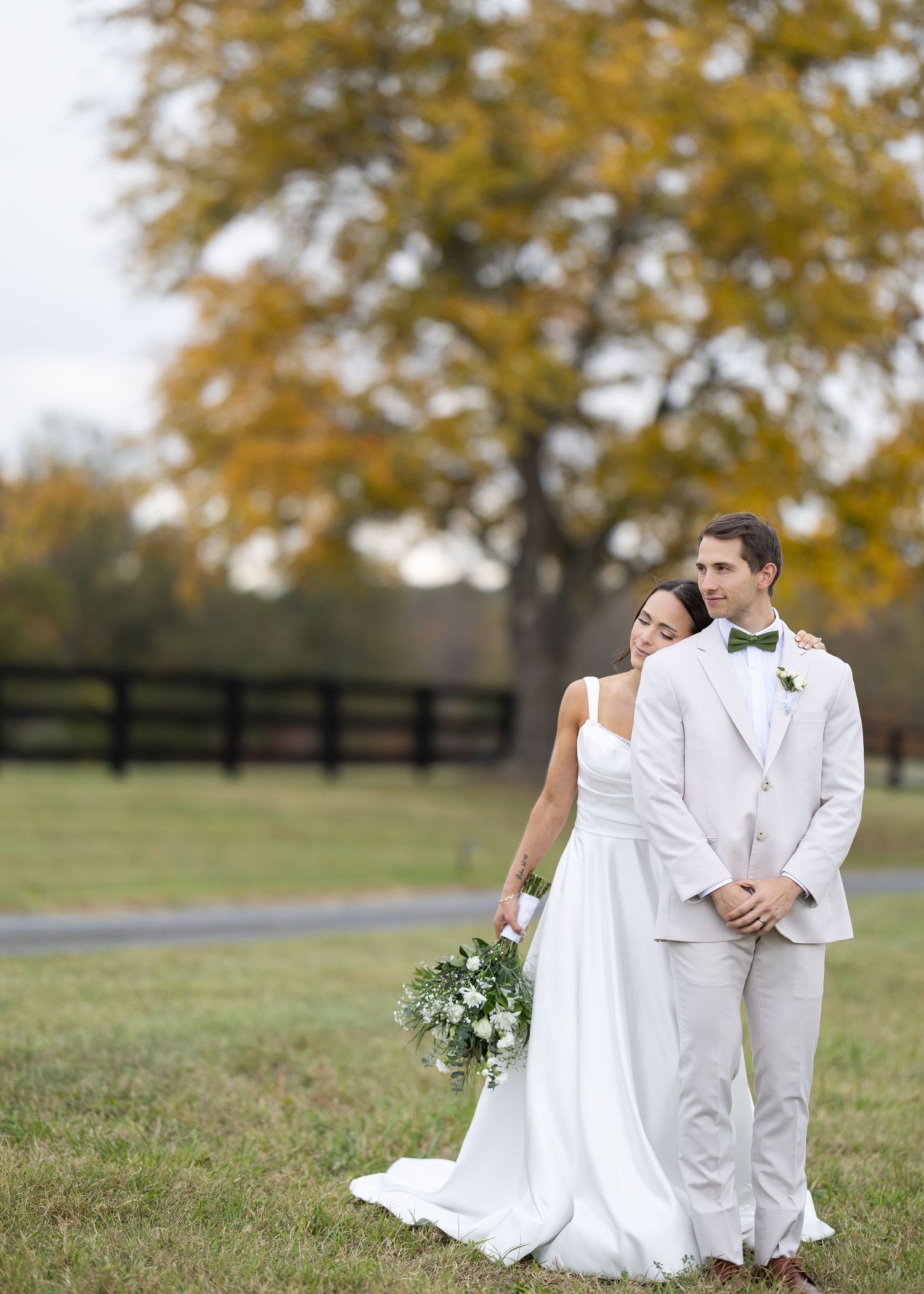 Bride and groom pose together in a green pasture with a vibrant autumn tree and wooden fence behind them.