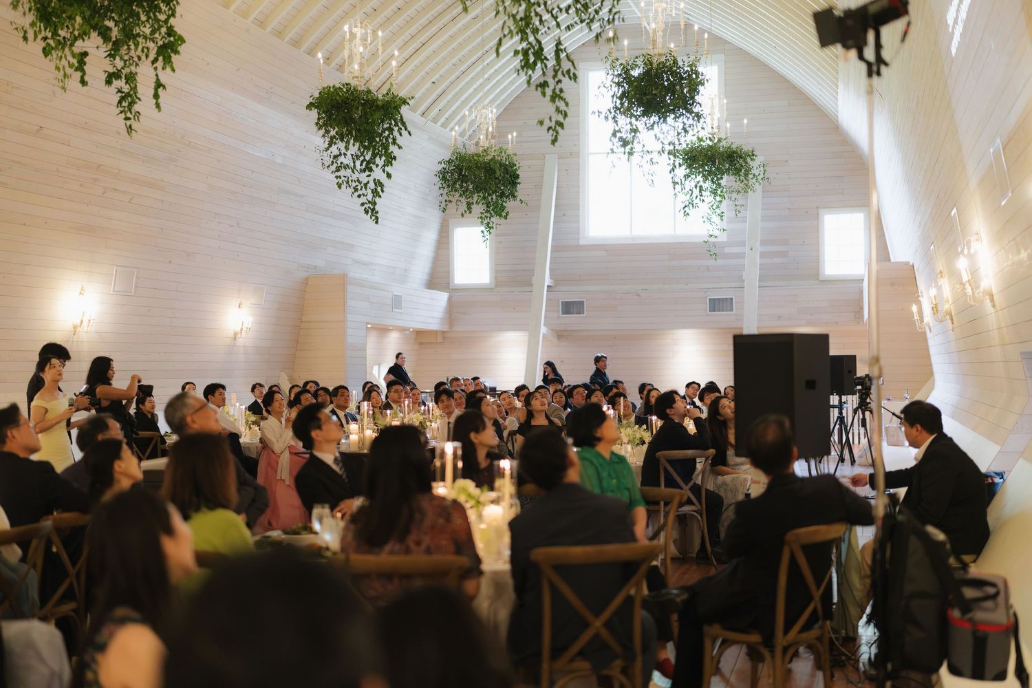 Elegant wedding reception in a white barn venue with hanging greenery, chandeliers, and guests seated at decorated tables.