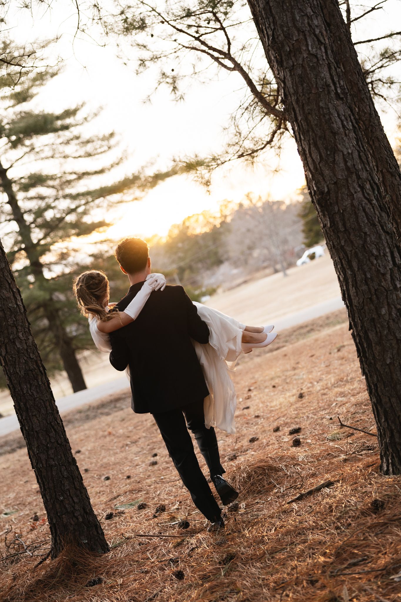 Groom carries bride in white gown through pine forest at golden sunset in romantic wedding portrait.