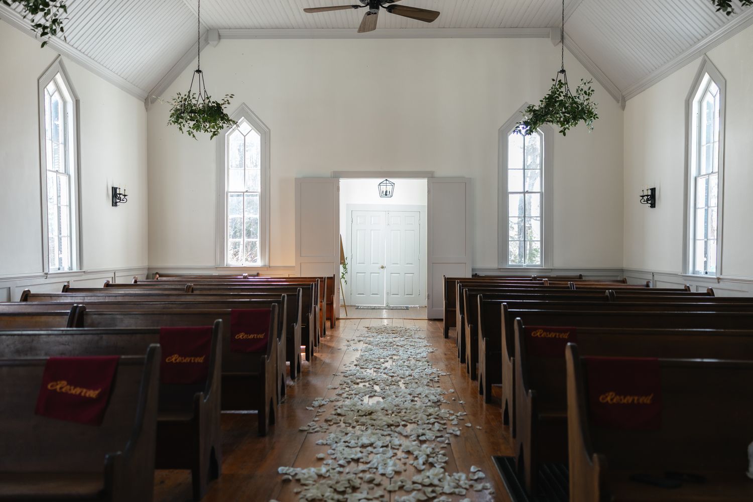 White chapel interior with wooden pews, petal-lined aisle, and hanging greenery decorations.