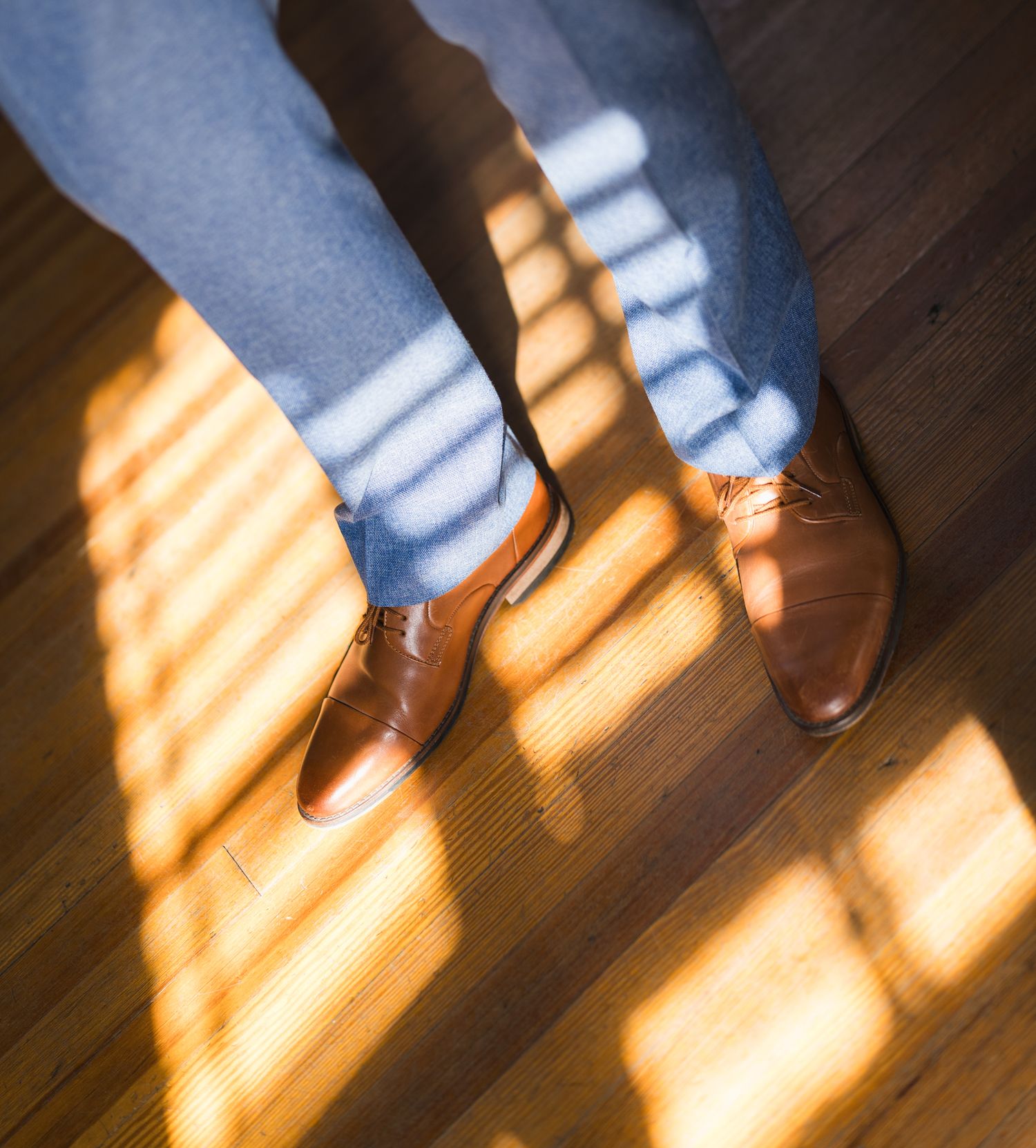 Close-up of groom wearing brown leather oxford shoes and blue jeans standing on a sunlit hardwood floor.