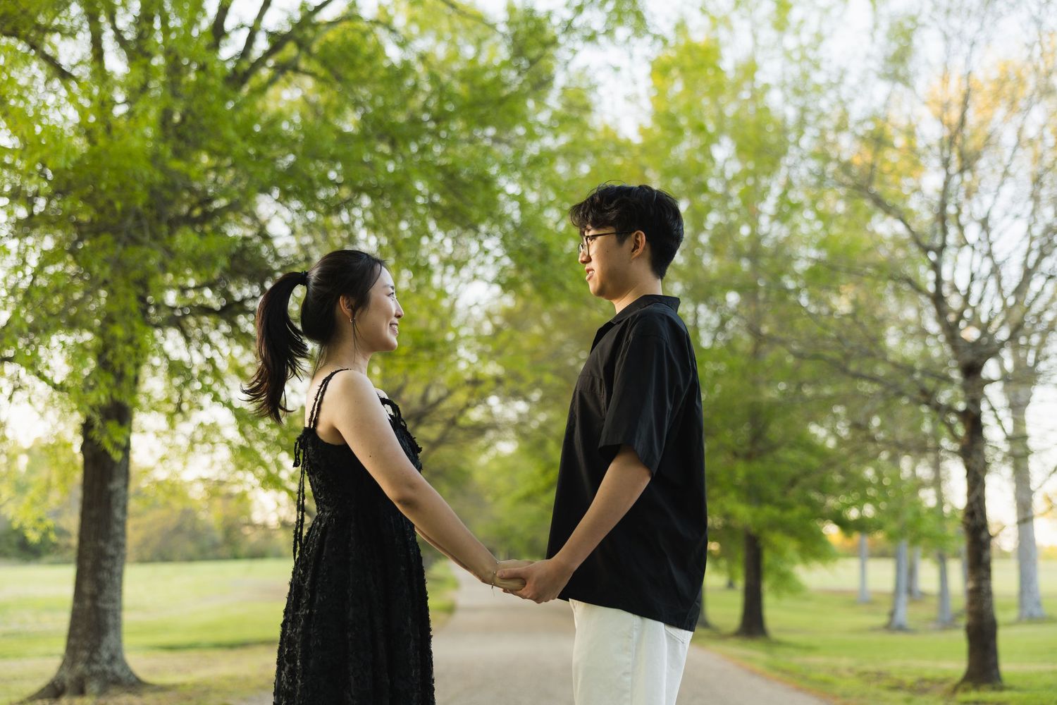 A young couple holds hands facing each other on a scenic tree-lined path in a park at golden hour.