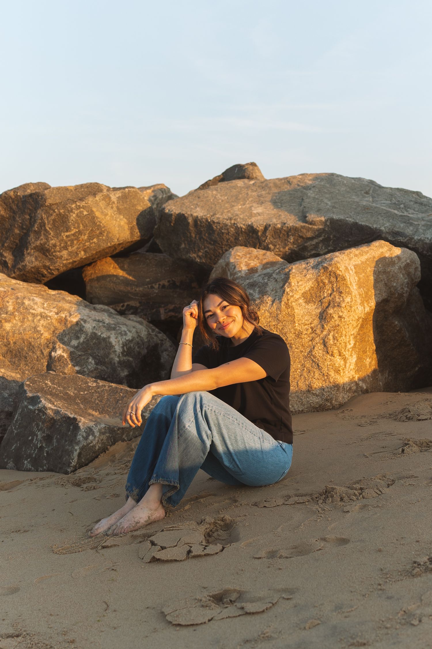 Smiling young woman in black tee and jeans sitting barefoot on sandy beach beside large golden rocks.