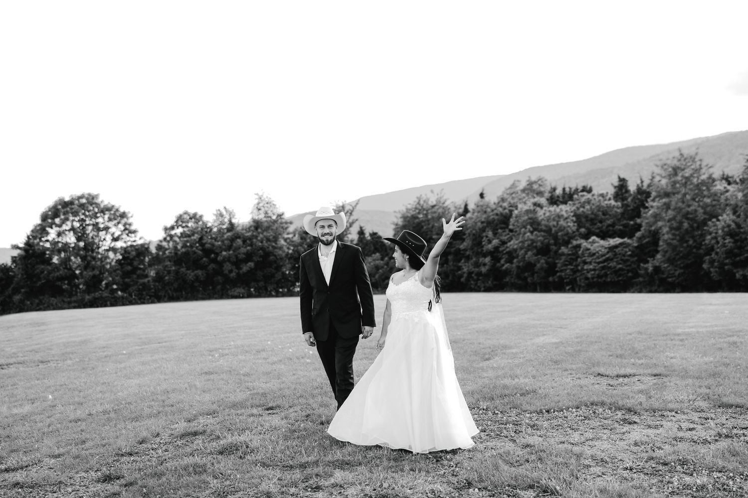 Black and white photo of a bride and groom holding hands walking across a grassy field, bride raising her arm in joy.