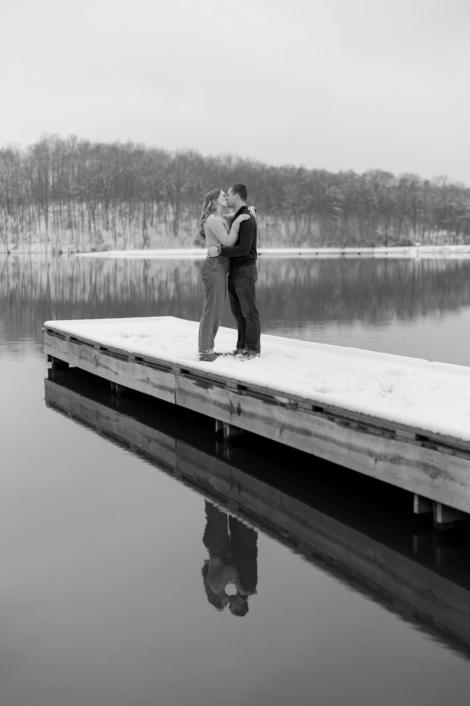 Black and white photo of couple kissing on a snowy dock reflected in a calm winter lake.
