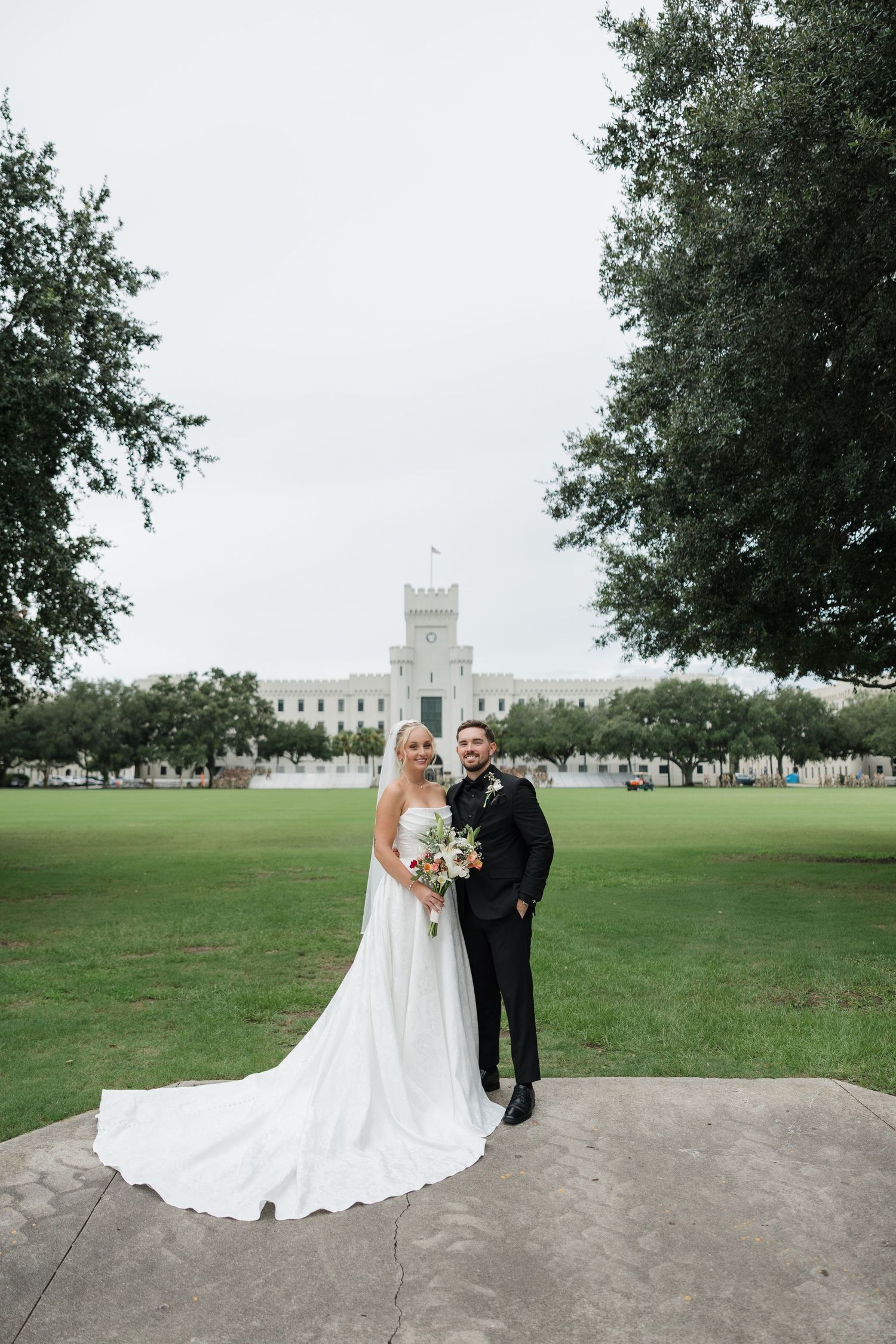 Bride and groom posing on green lawn in front of a white castle-like building on their wedding day.