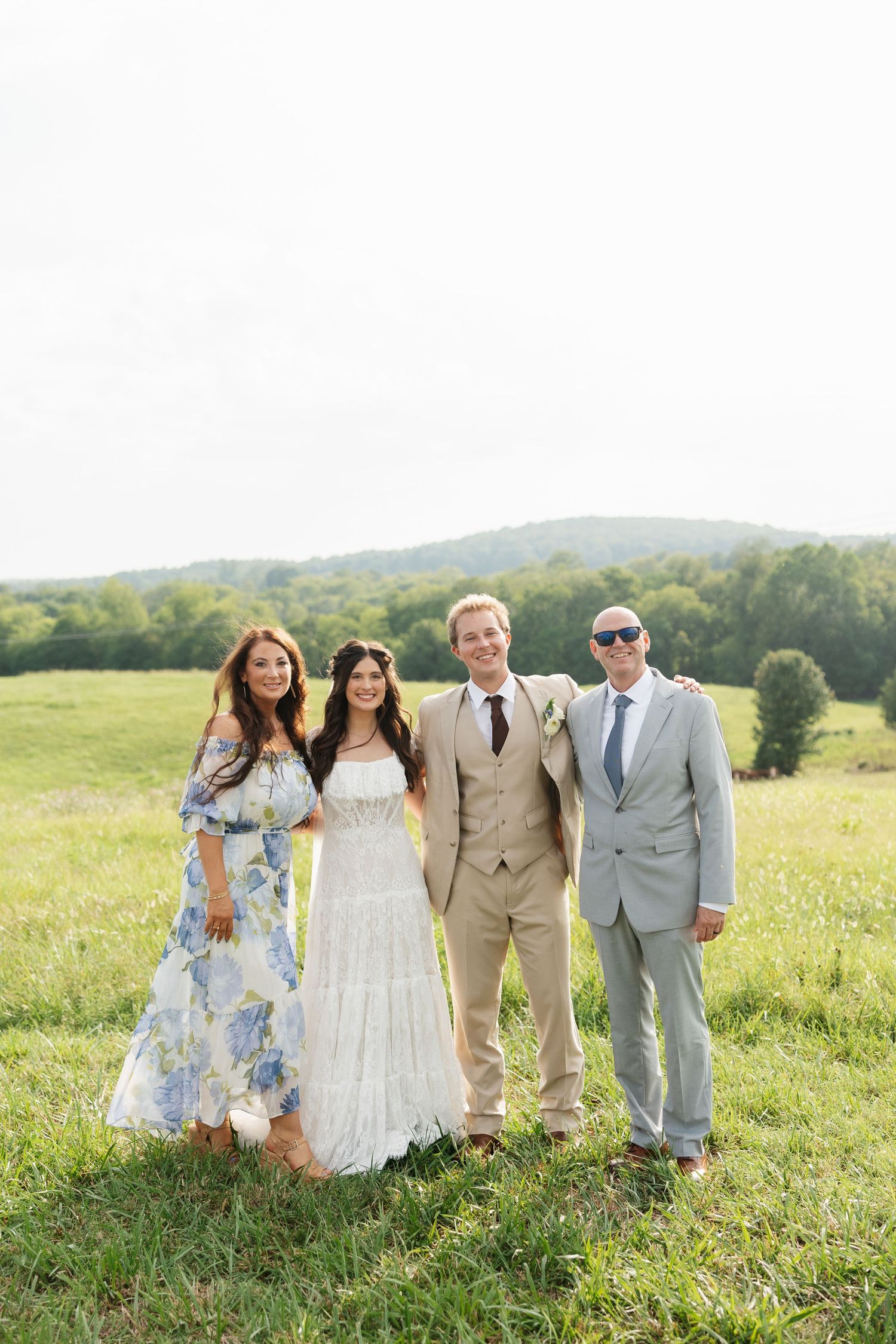 Wedding party of four posing in a green meadow, bride in white dress, groom in tan suit, guests in floral and gray attire.