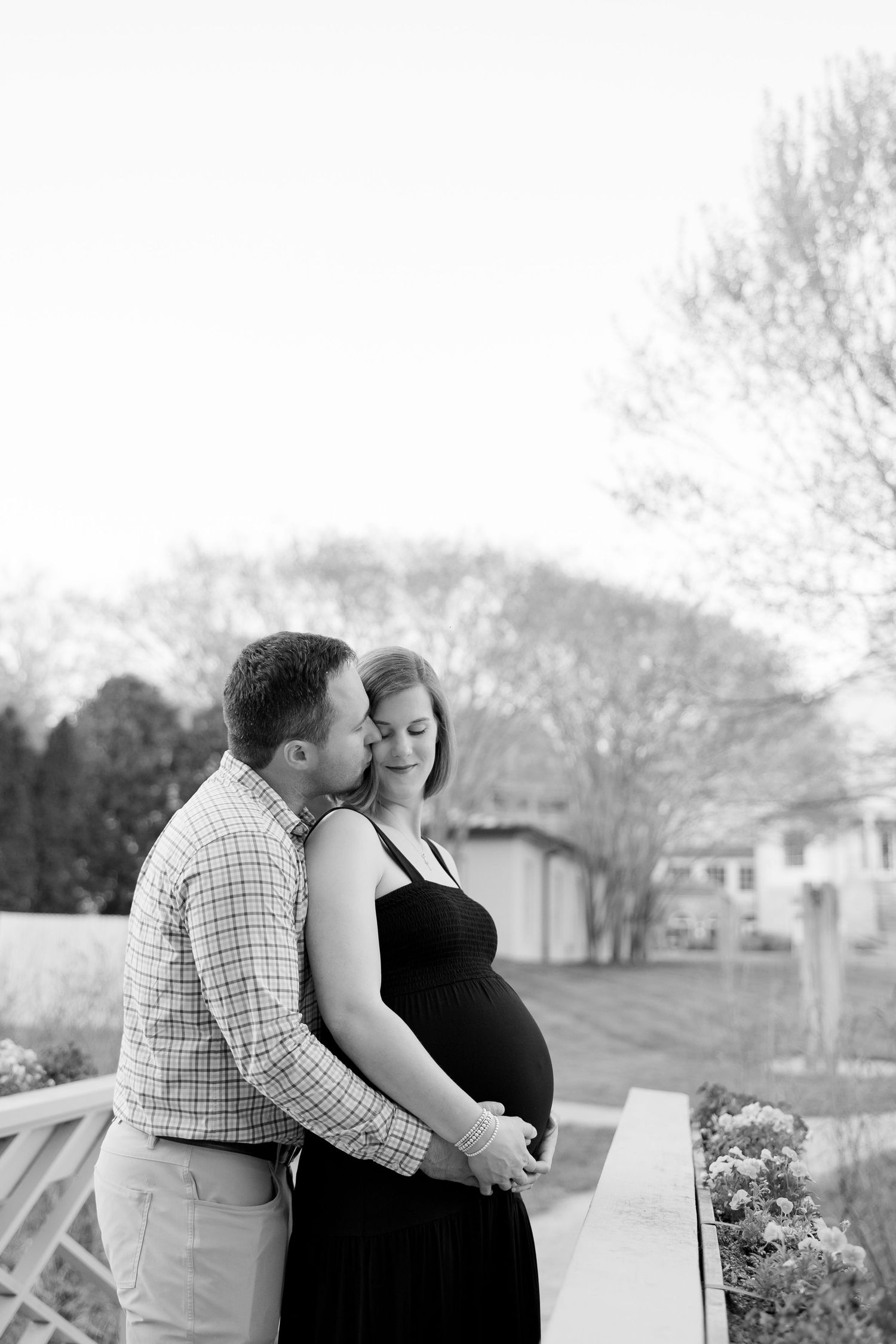 Black and white maternity photo of a couple outdoors, man kissing pregnant woman's cheek while cradling her baby bump.