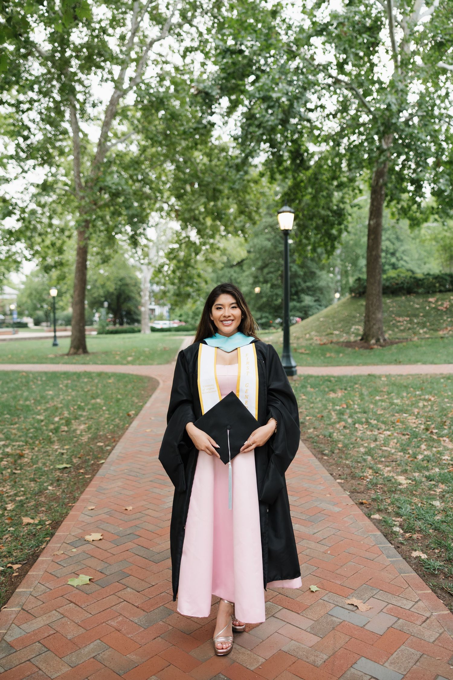 Smiling graduate in black gown and pink dress holds cap on brick pathway surrounded by green trees.