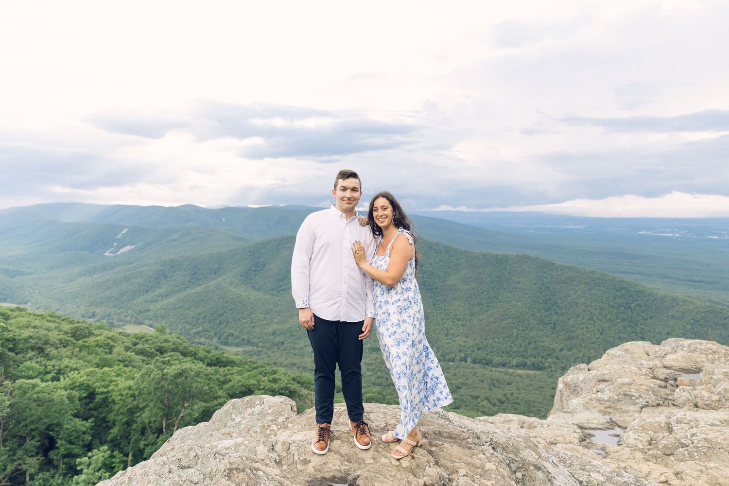 A happy couple poses on a rocky overlook with stunning panoramic mountain views and cloudy skies behind them.