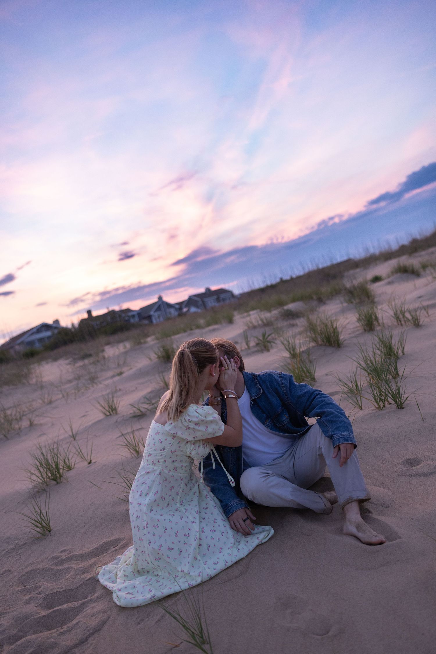 Romantic couple sitting together on sandy beach dunes under a colorful purple and pink sunset sky.