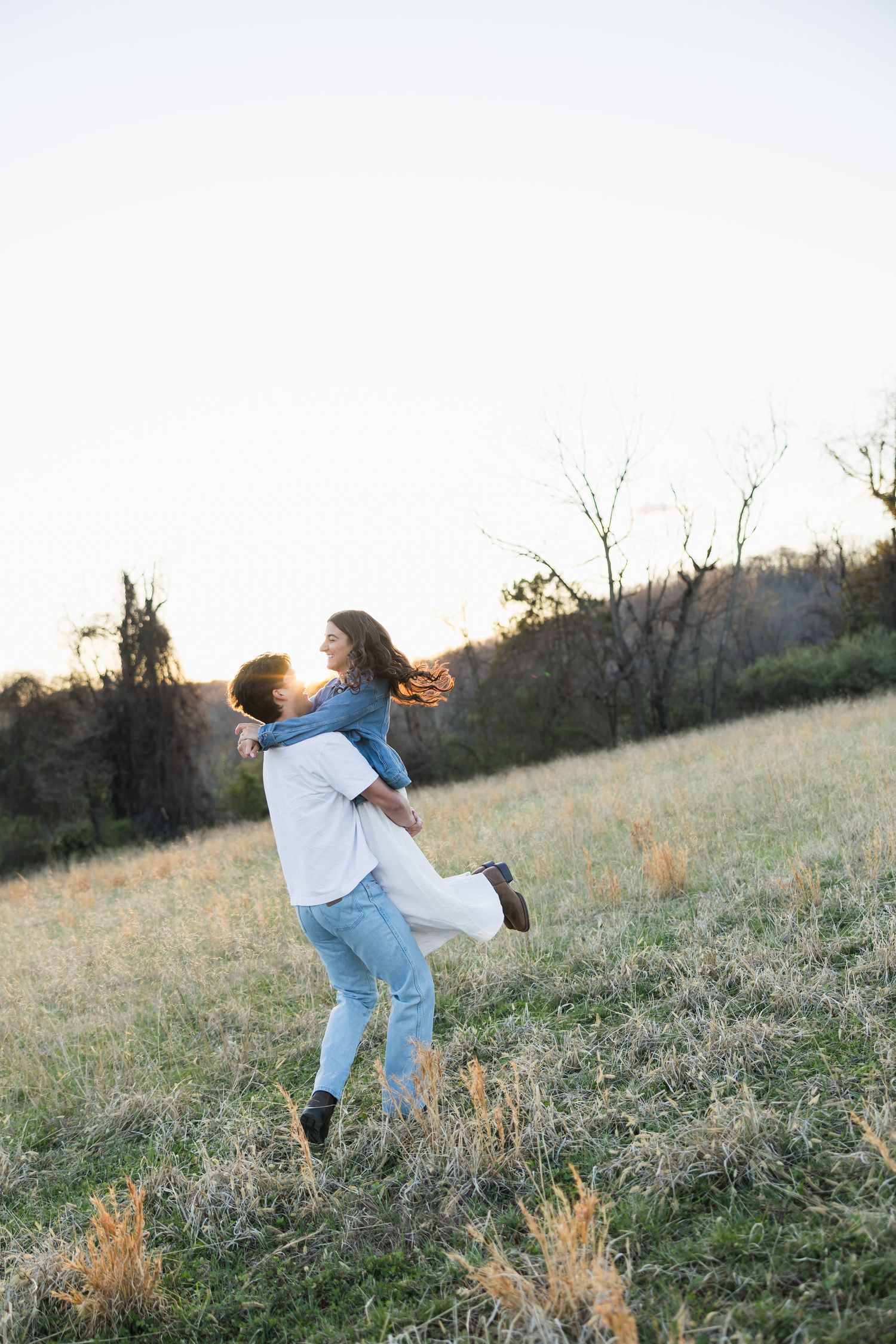 A joyful couple spinning in a golden field at sunset, the man lifting the woman in a white dress and denim jacket.