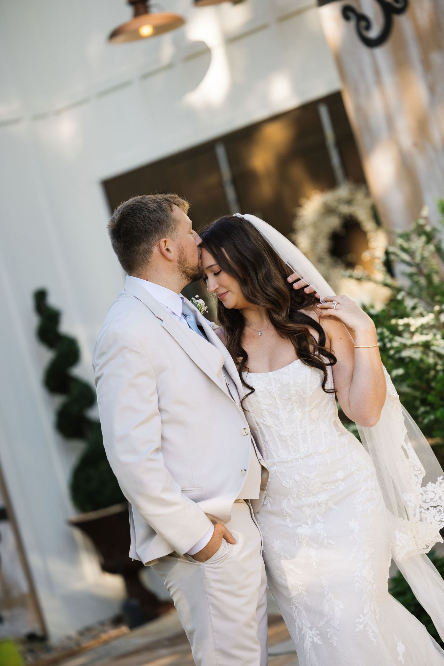 Bride and groom share a tender moment, groom in beige suit kissing bride's forehead in white lace gown with veil.