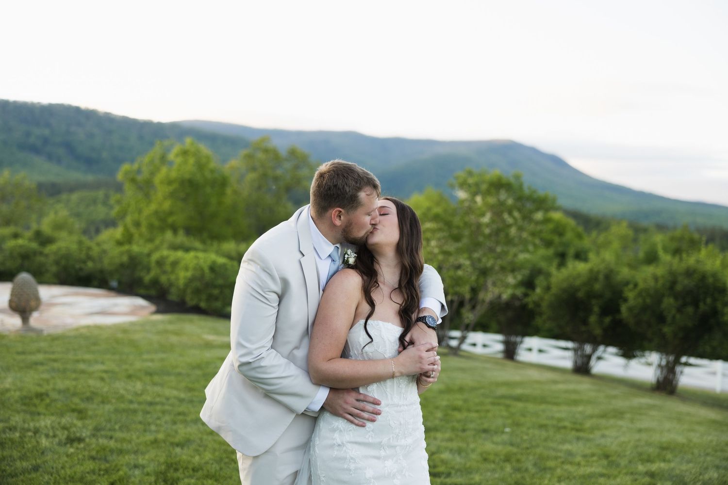 Bride and groom sharing a kiss outdoors with lush green mountains and trees in the background.