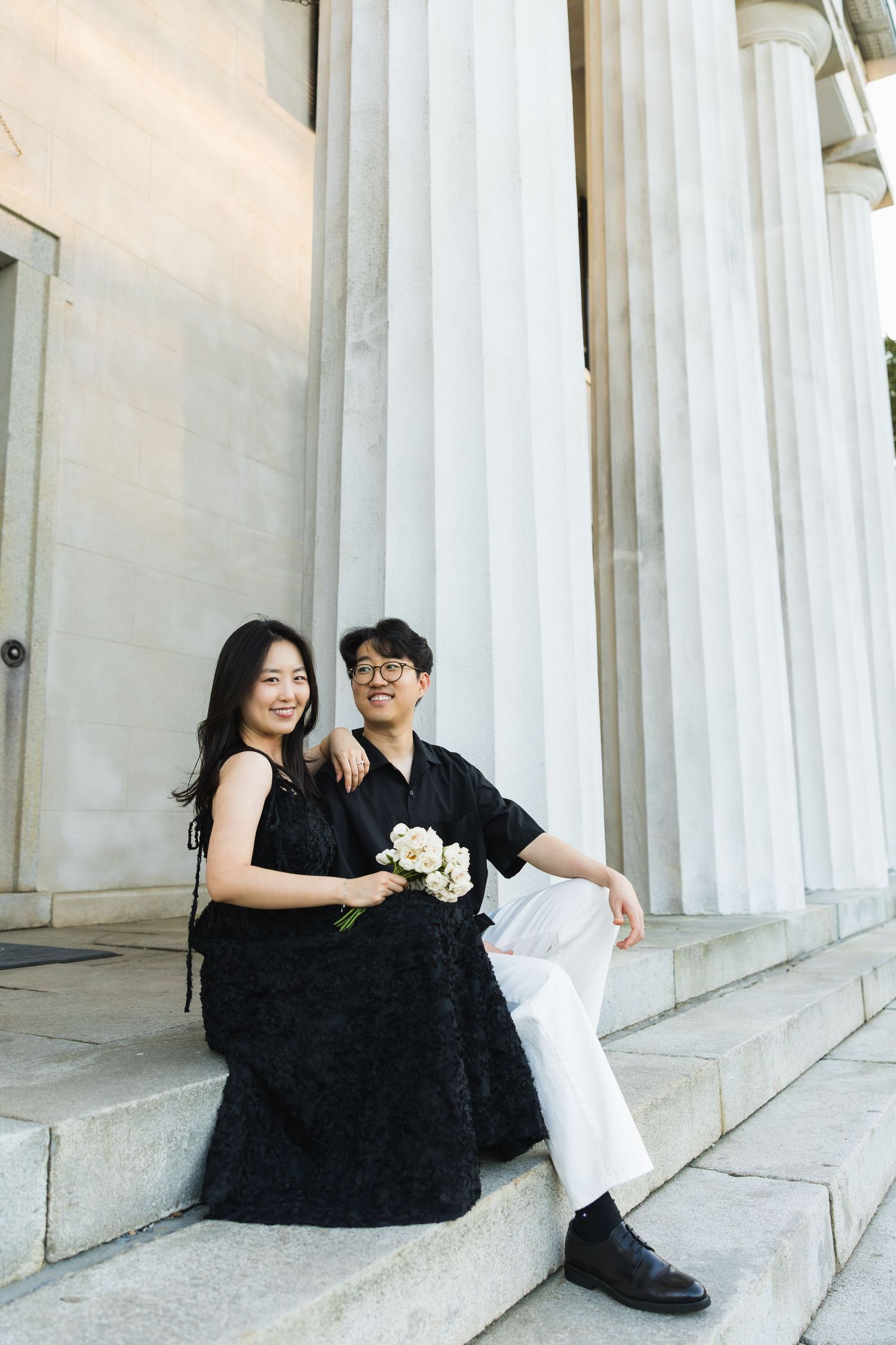A couple in matching black and white outfits sits on stone steps with white columns, the woman holding white flowers.