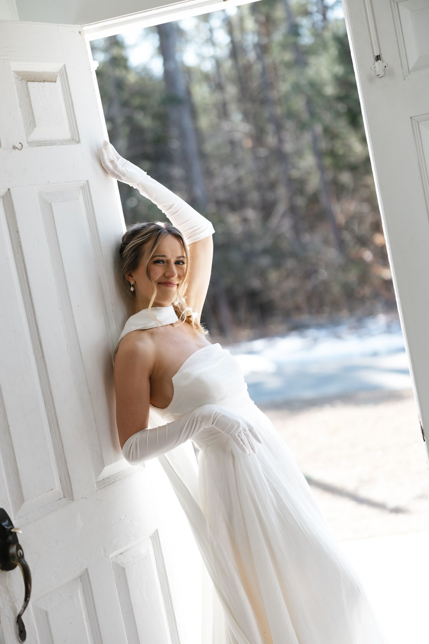 Bride in backless white gown and gloves posing gracefully in open doorway with snowy backdrop.