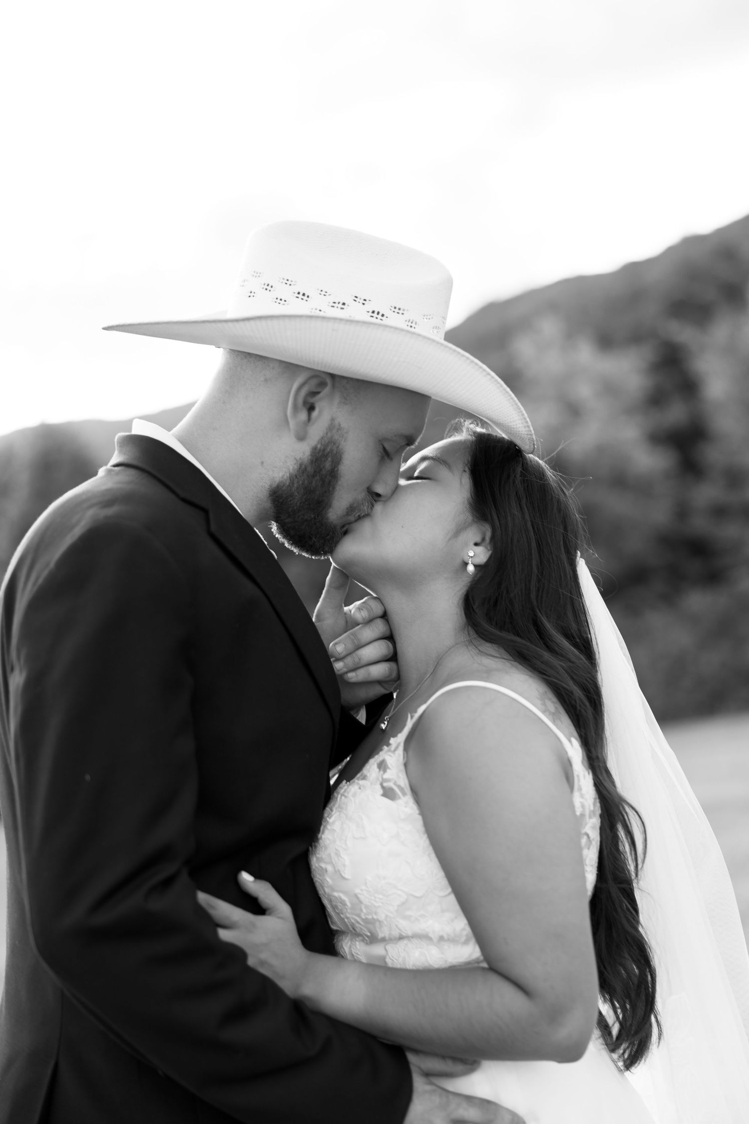 Black and white photo of a bride and groom sharing a kiss, groom wearing a cowboy hat, outdoors with mountains behind.