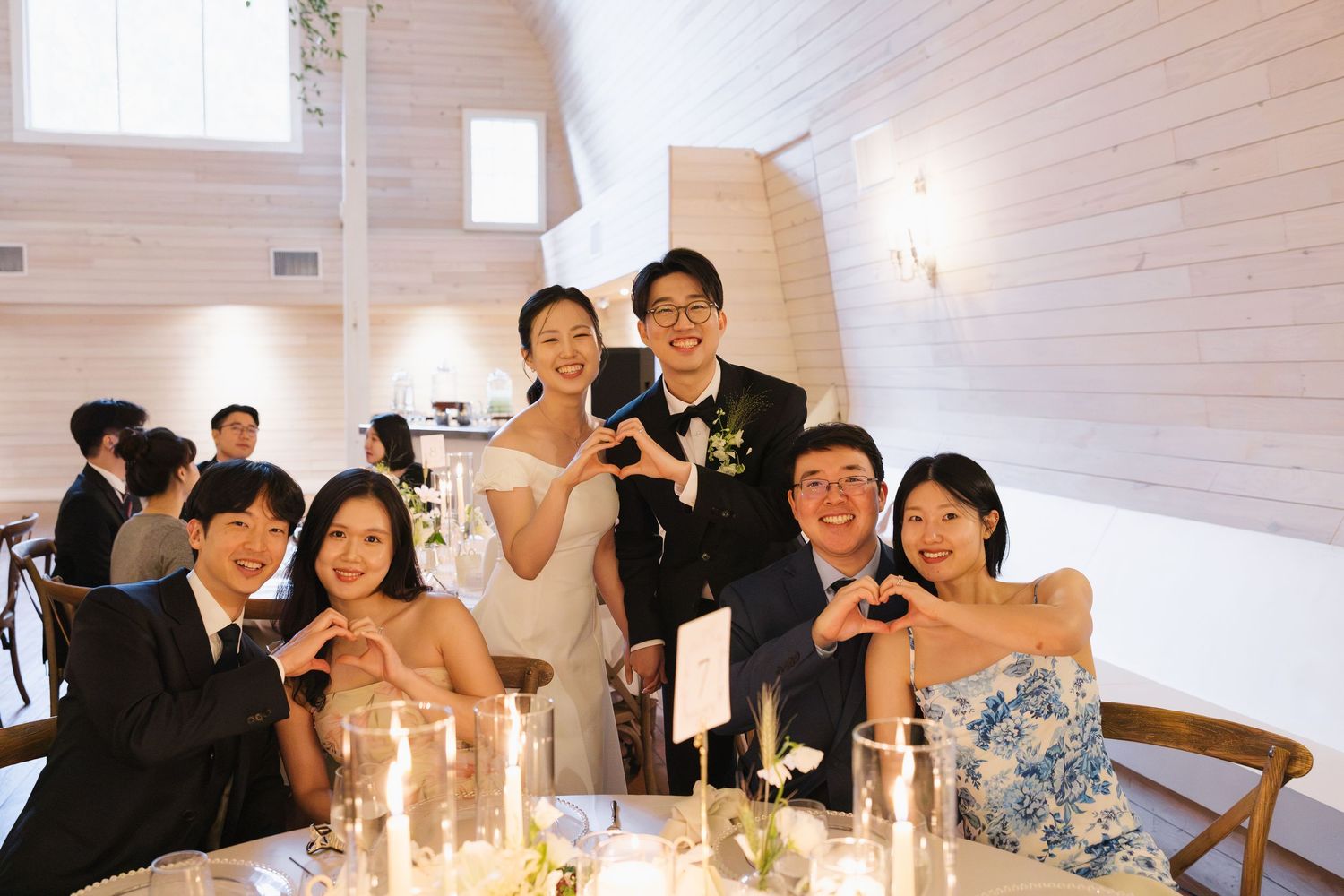 Group of smiling wedding guests making heart gestures with hands, posing with bride and groom at reception table.