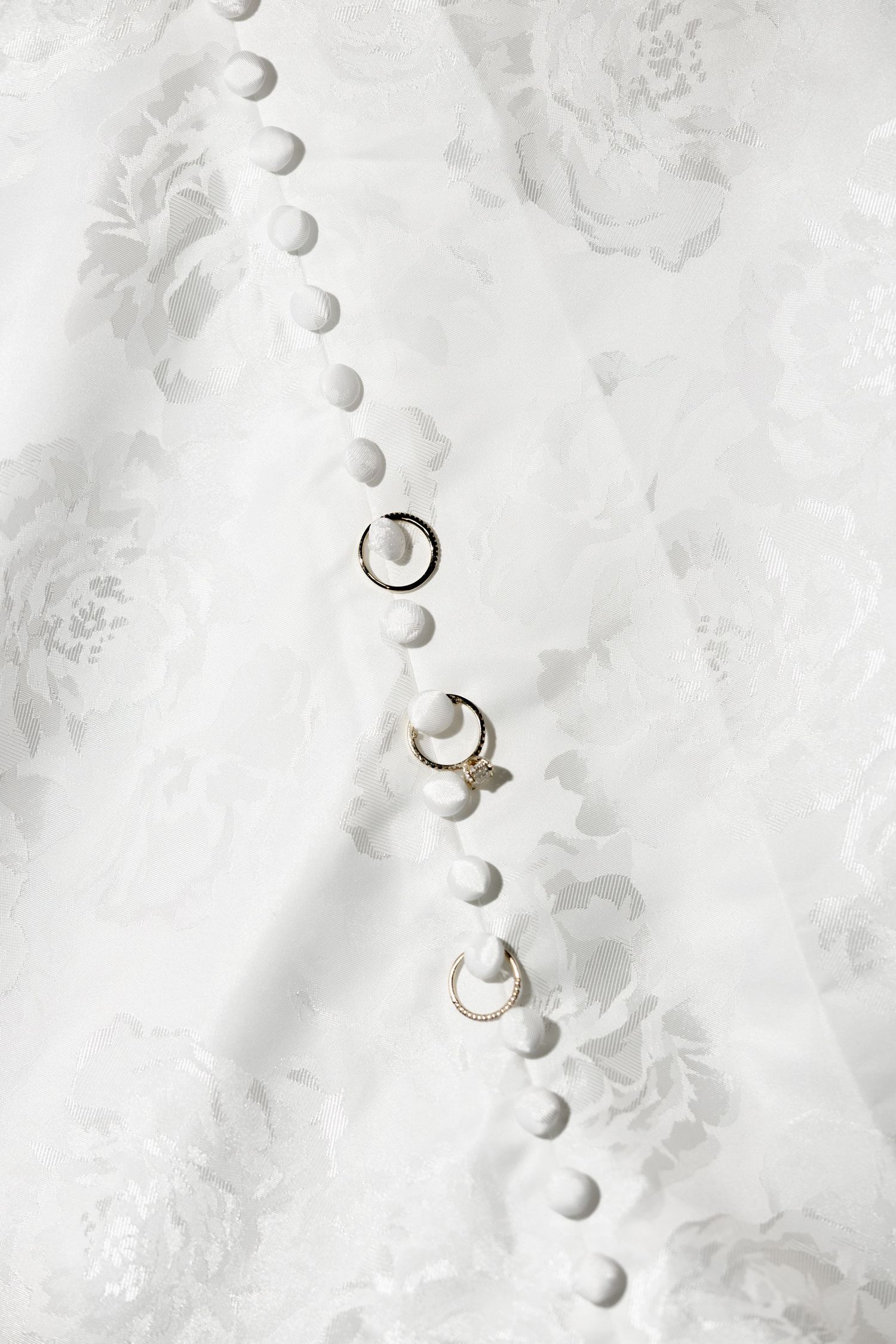 Gold wedding rings and earrings displayed on white floral lace bridal gown with pearl buttons.