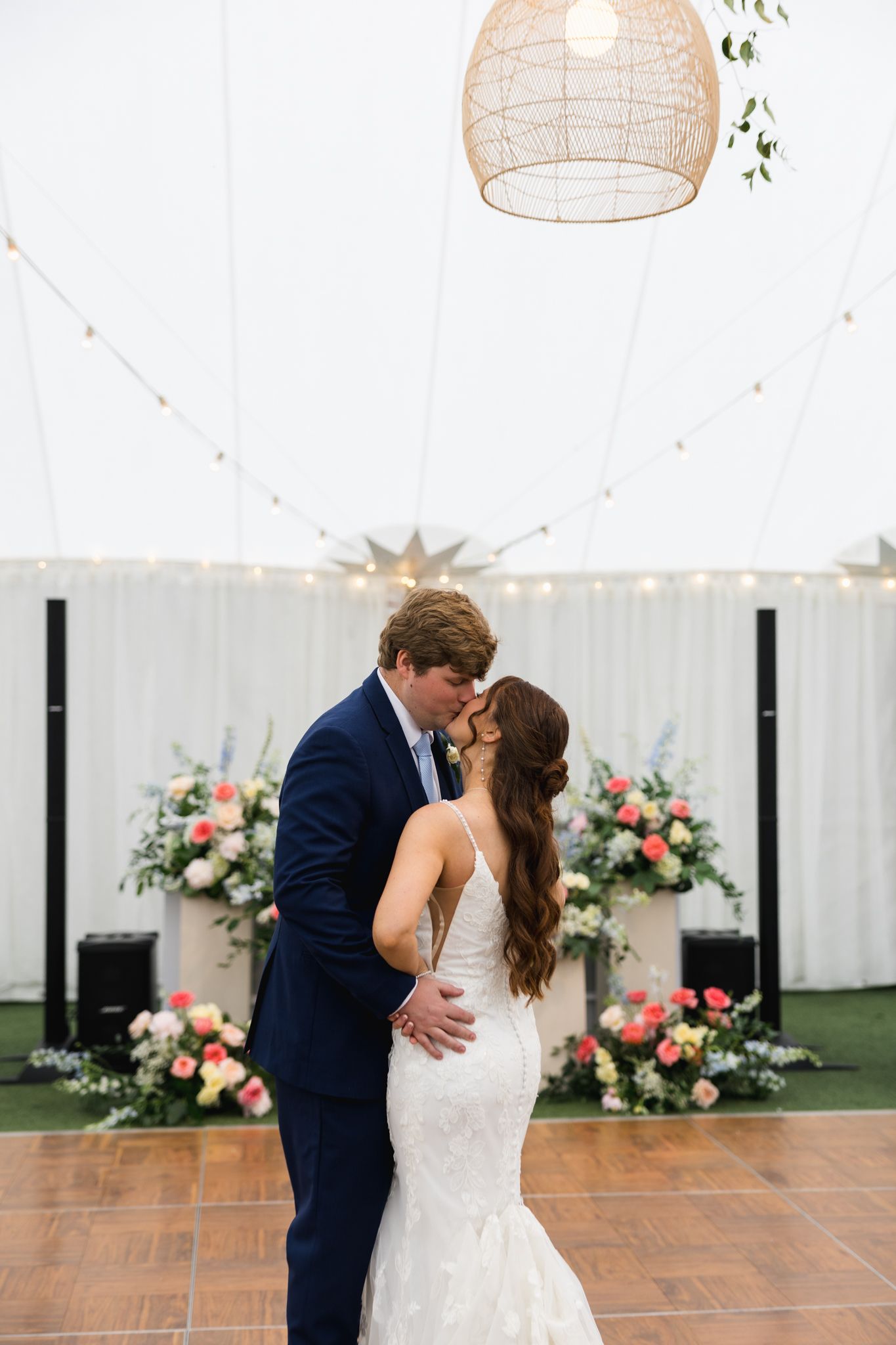 Newlyweds share first dance kiss under string lights inside elegant white wedding reception tent.