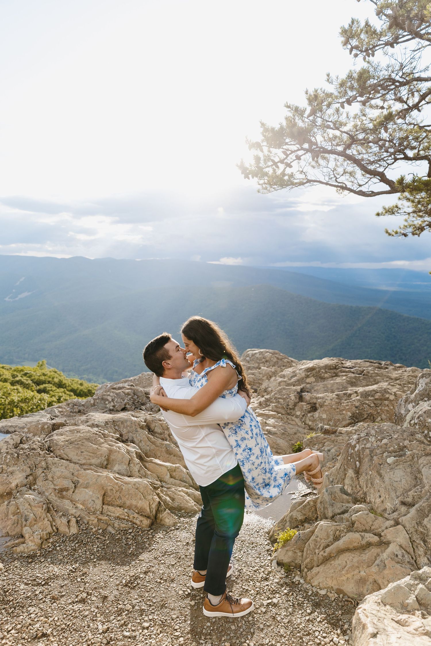 Man lifting woman in floral dress on rocky mountain summit in golden sunlight.