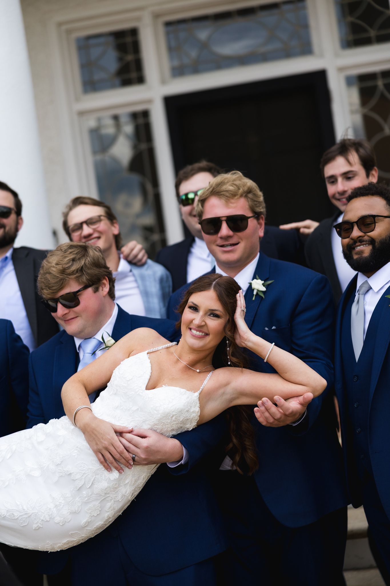 Groom lifts bride surrounded by groomsmen in navy suits and sunglasses, posing outdoors at a wedding celebration.