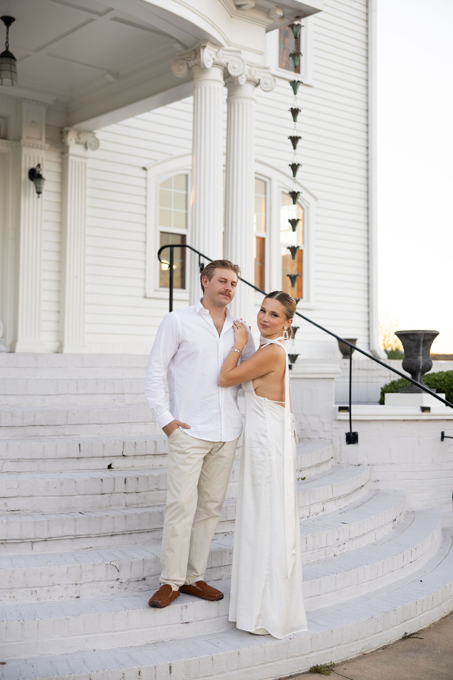 Elegant couple in white attire posing on steps of a grand white colonial building with tall columns.