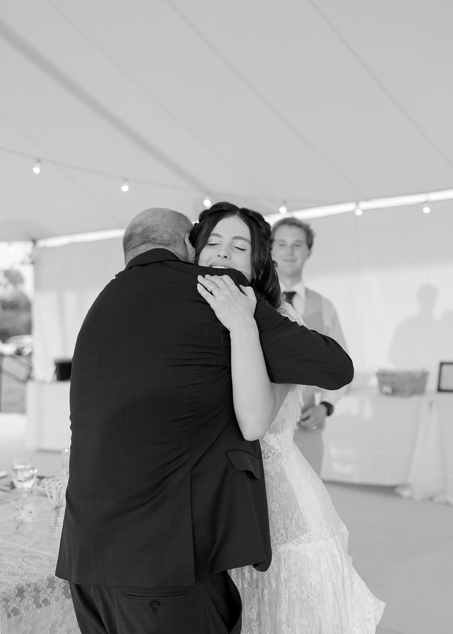 Bride in white wedding dress sharing emotional hug with guest under tent at reception.