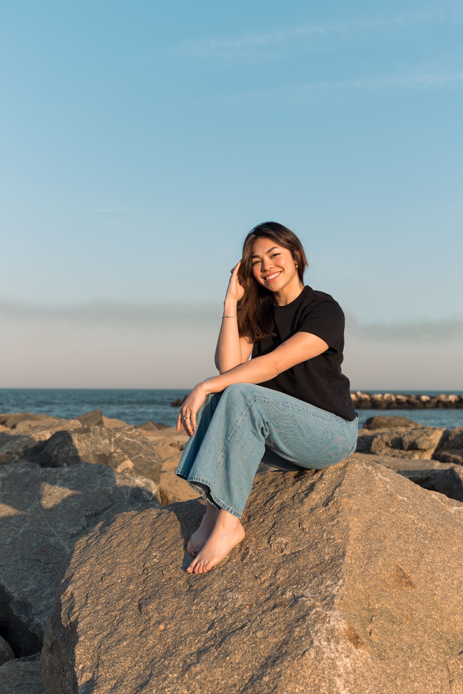 Happy young woman in black t-shirt and jeans sitting barefoot on rocks by the ocean at sunset.