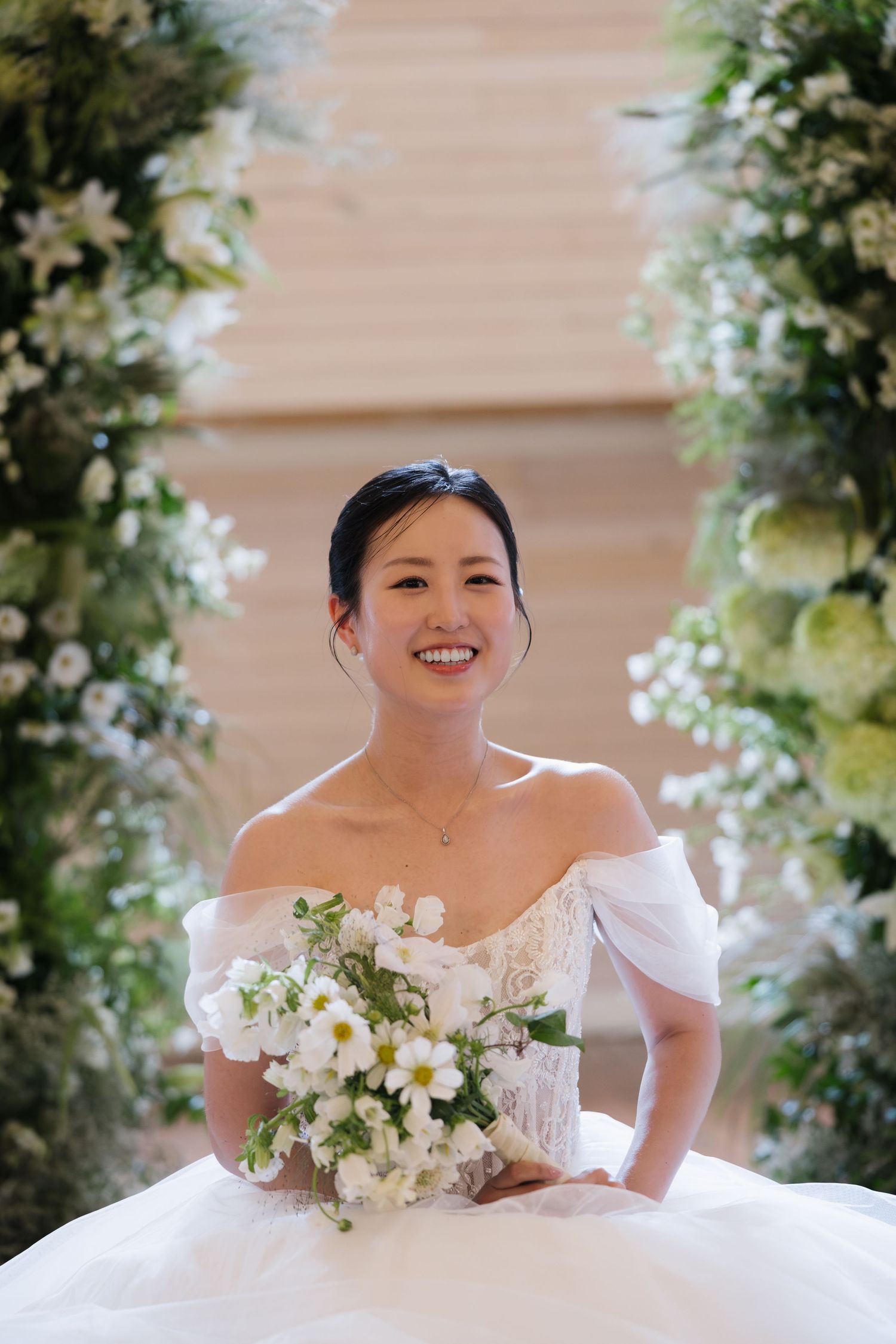 Smiling bride in off-shoulder white gown holds white floral bouquet surrounded by lush greenery.