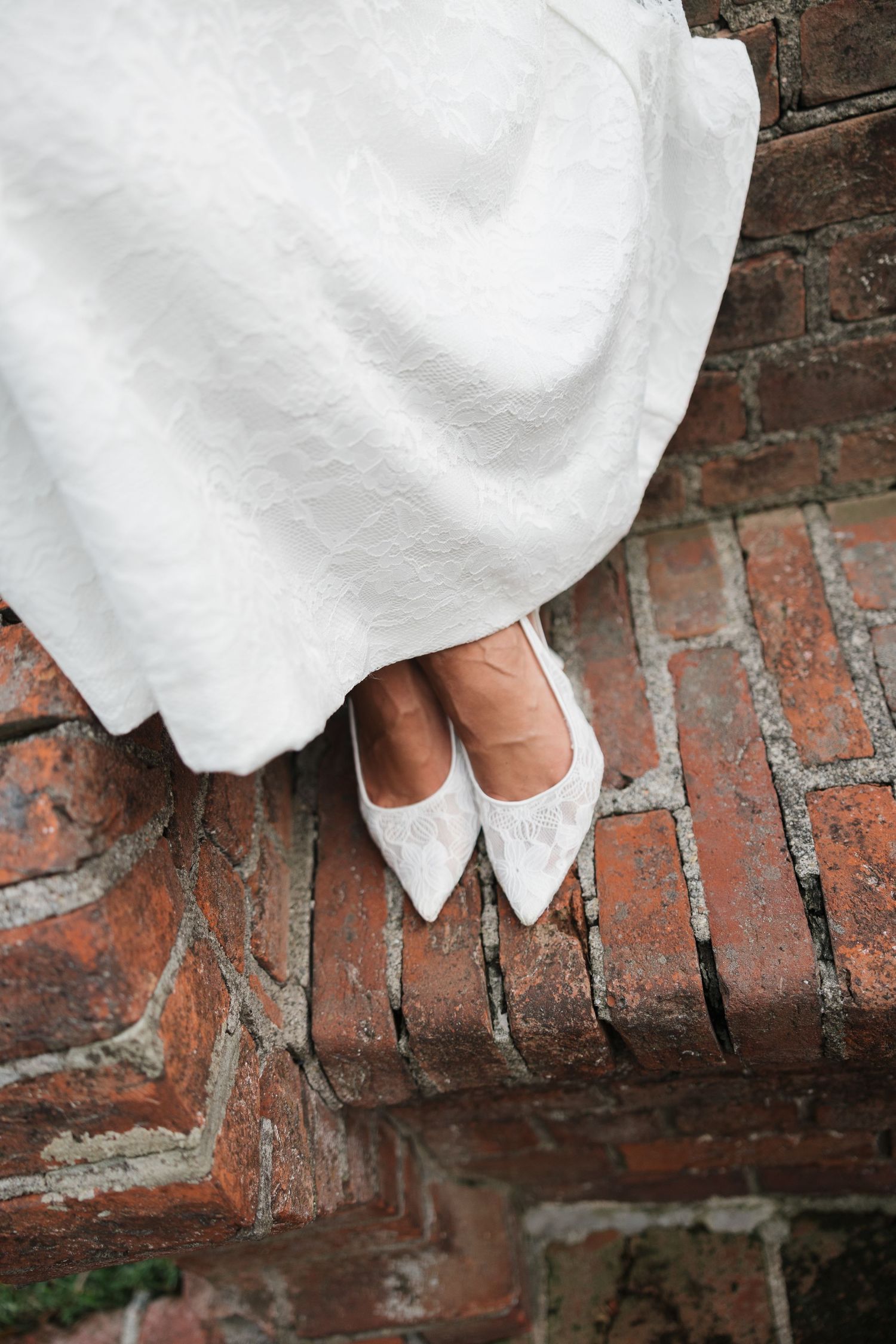Bride wearing white lace pointed-toe flats peeking beneath a white wedding gown on a rustic red brick surface.